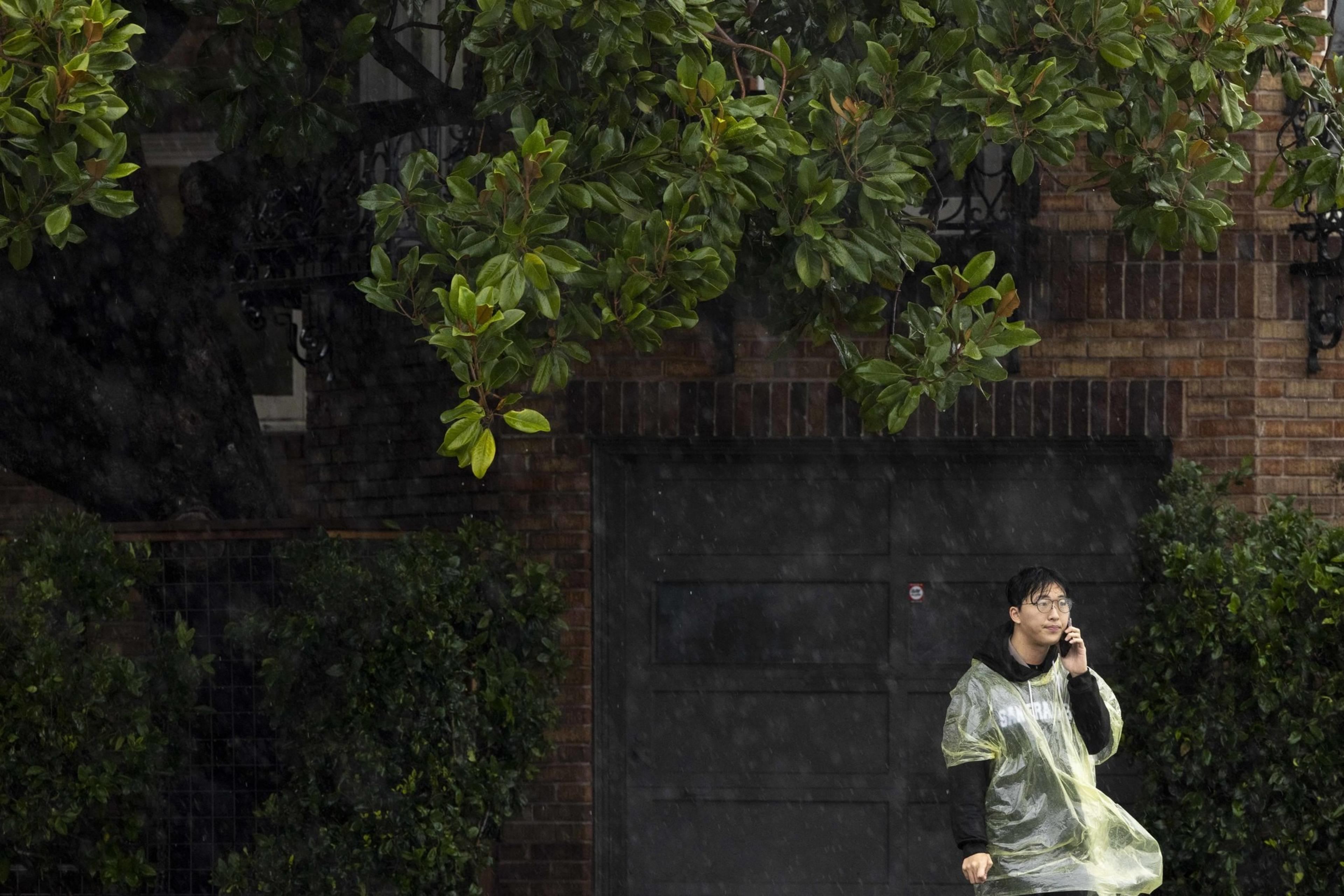 A person in a yellow rain poncho stands under a tree during a rain shower, talking on a phone. They're beside a brick building with a dark garage door.