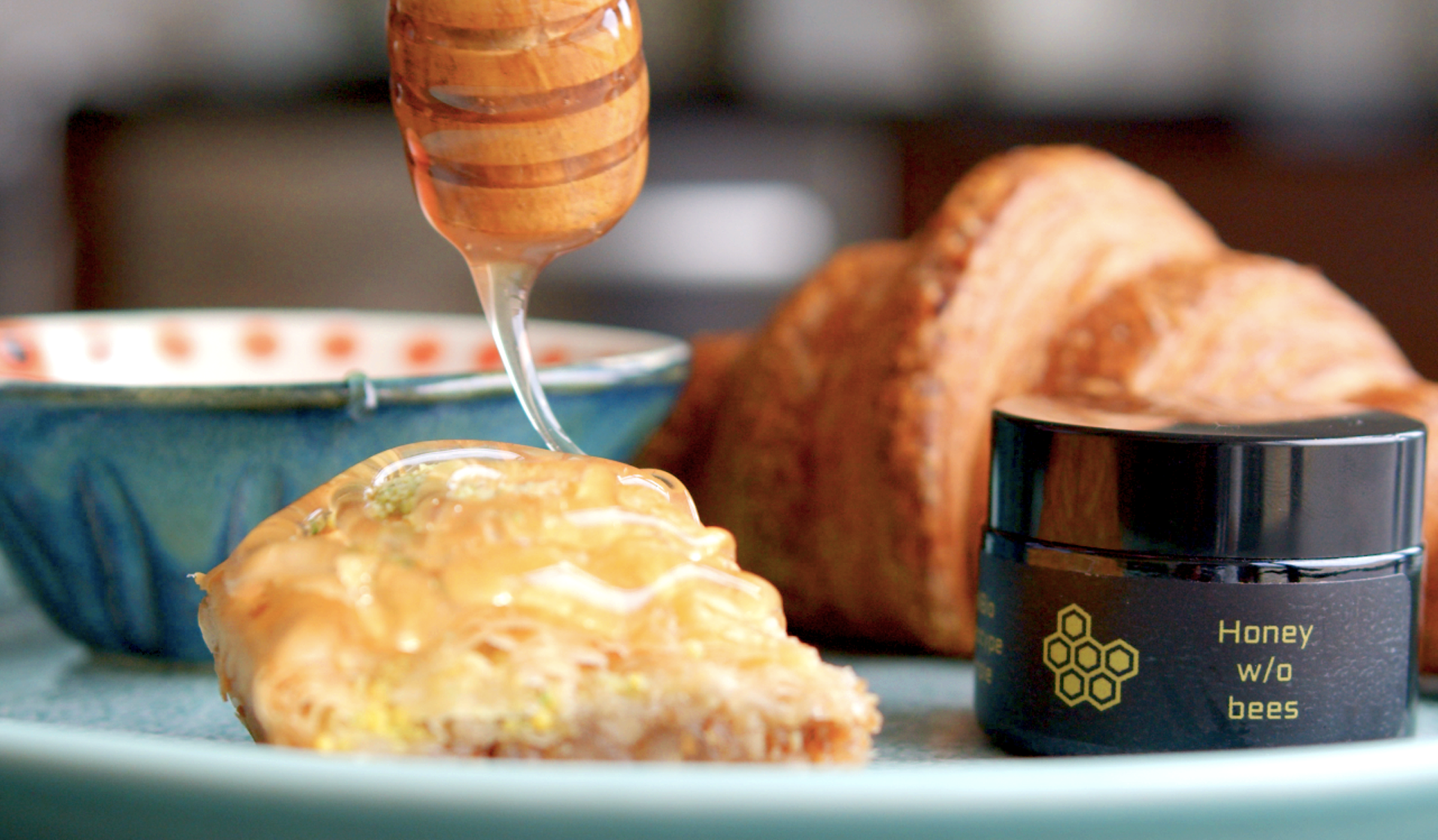 Honey is being drizzled onto a pastry on a plate, next to a croissant and a jar labeled "Honey w/o bees."