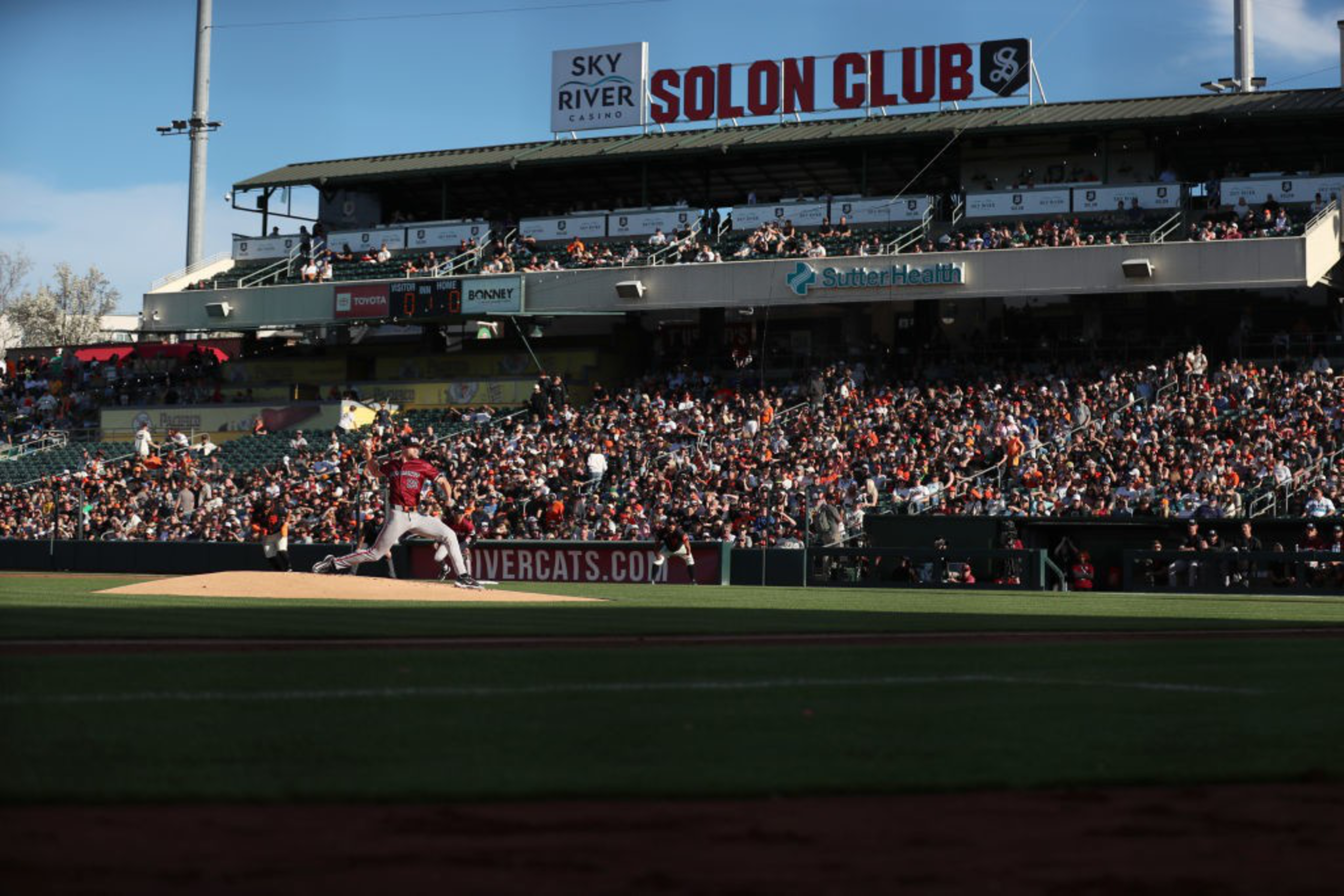 A baseball pitcher on the mound prepares to throw the ball in a packed stadium under a clear sky, with advertisements visible in the background.