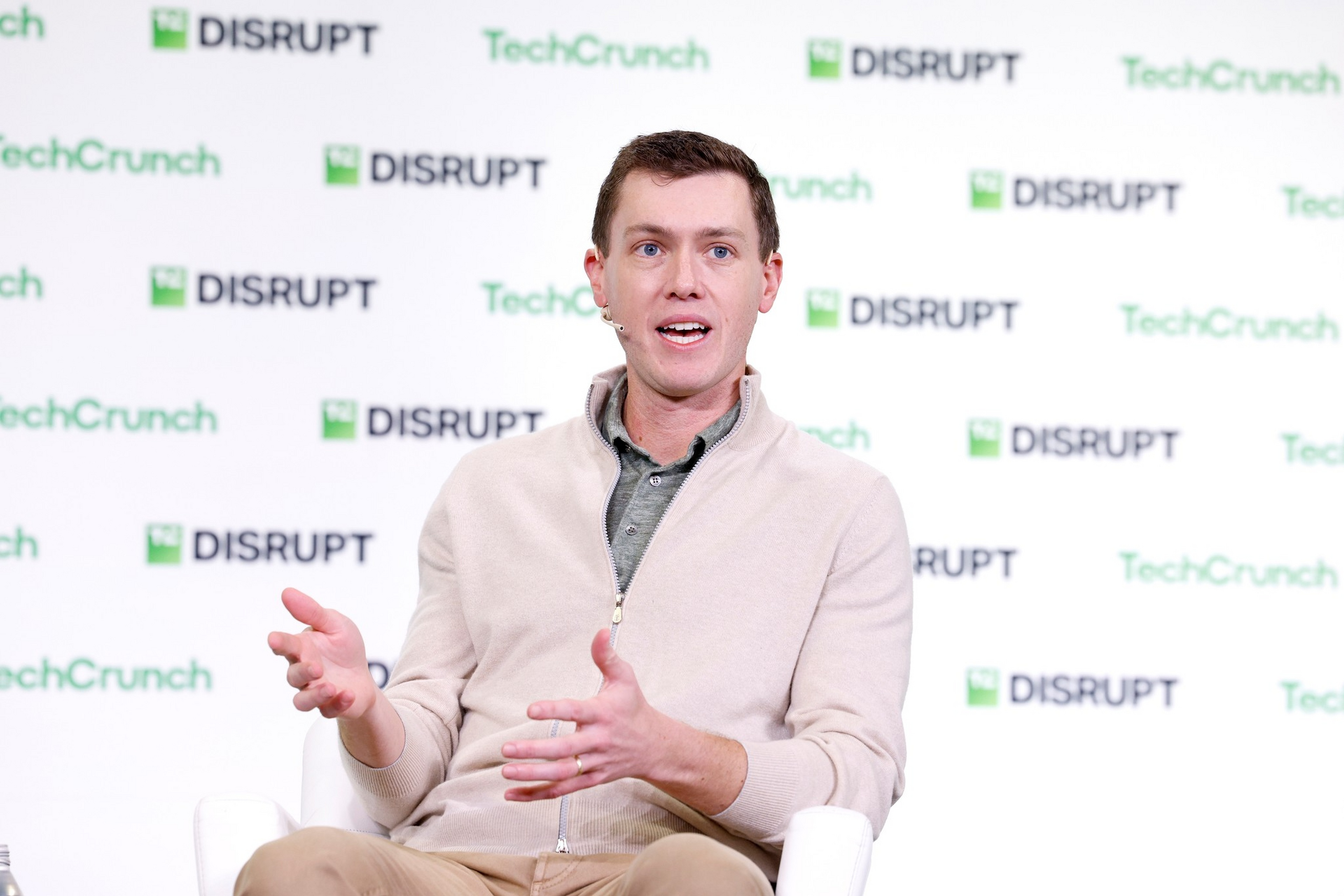 A man in a beige zip-up sweater and khaki pants gestures with his hands while speaking at a TechCrunch Disrupt event.
