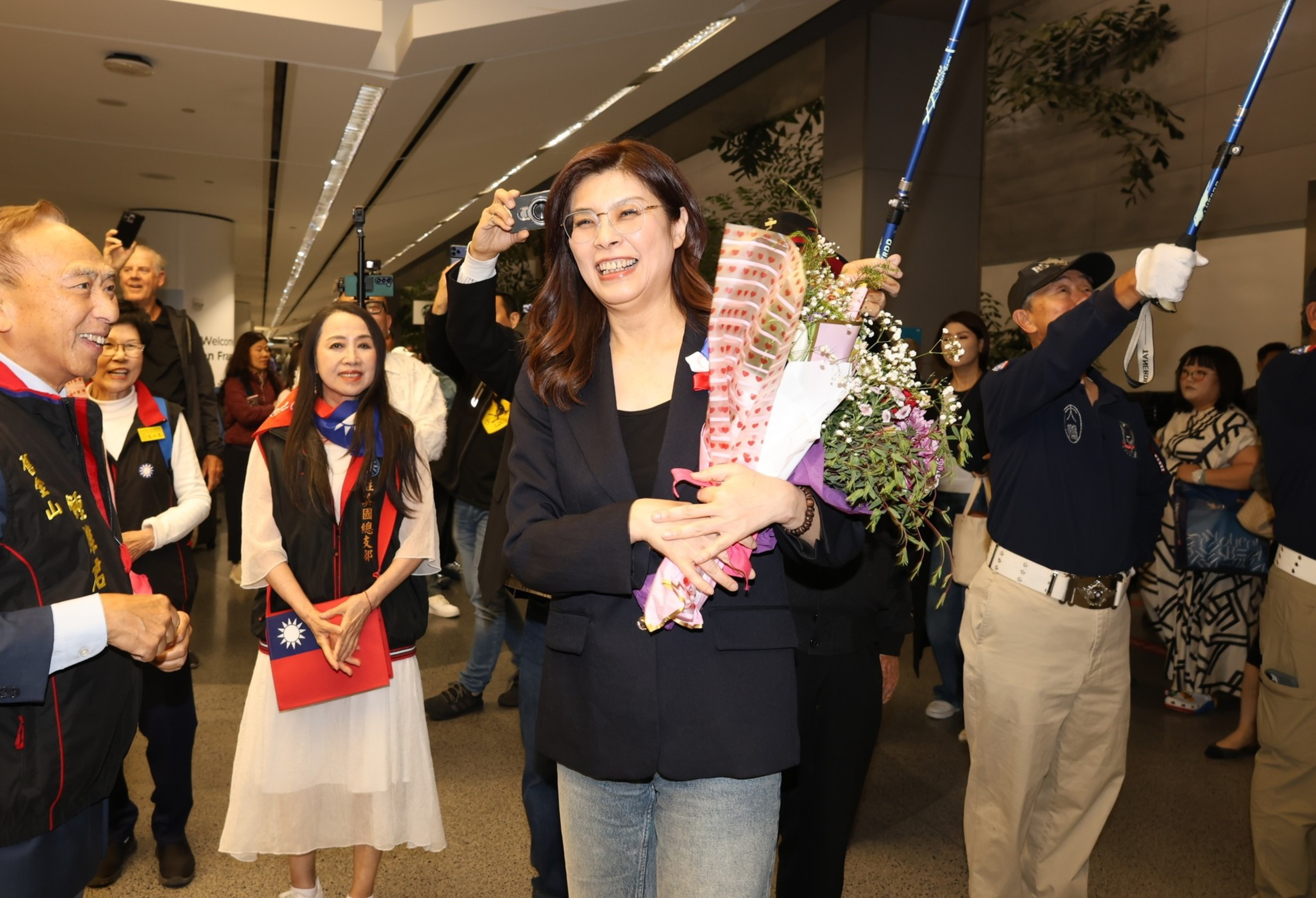 A smiling woman holding bouquets of flowers is surrounded by people, including uniformed officers holding swords raised in a ceremonial arch.