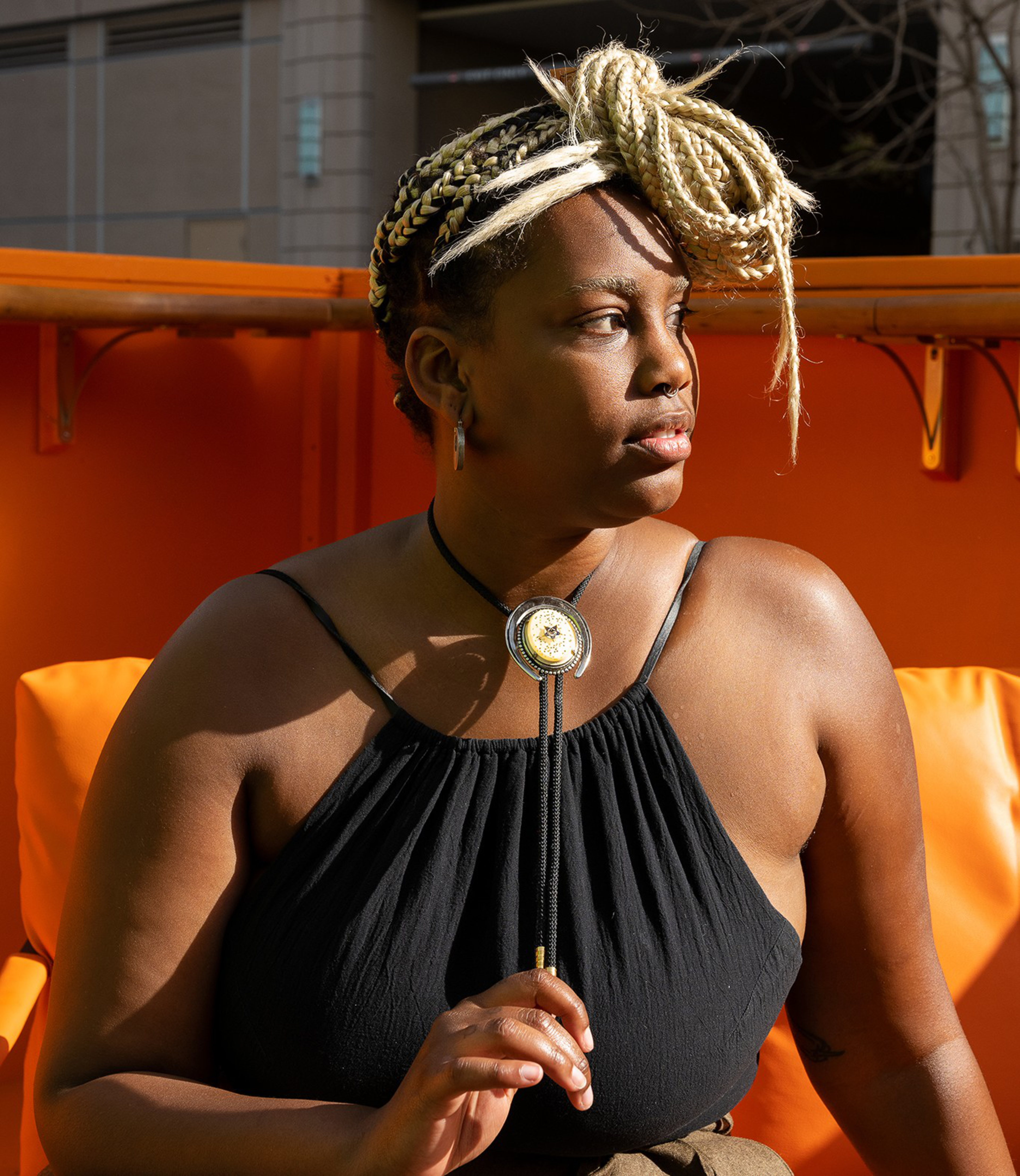 A woman with blonde braided hair styled in a loop wears a black sleeveless top and a bolo tie, sitting on an orange chair, looking to the side.