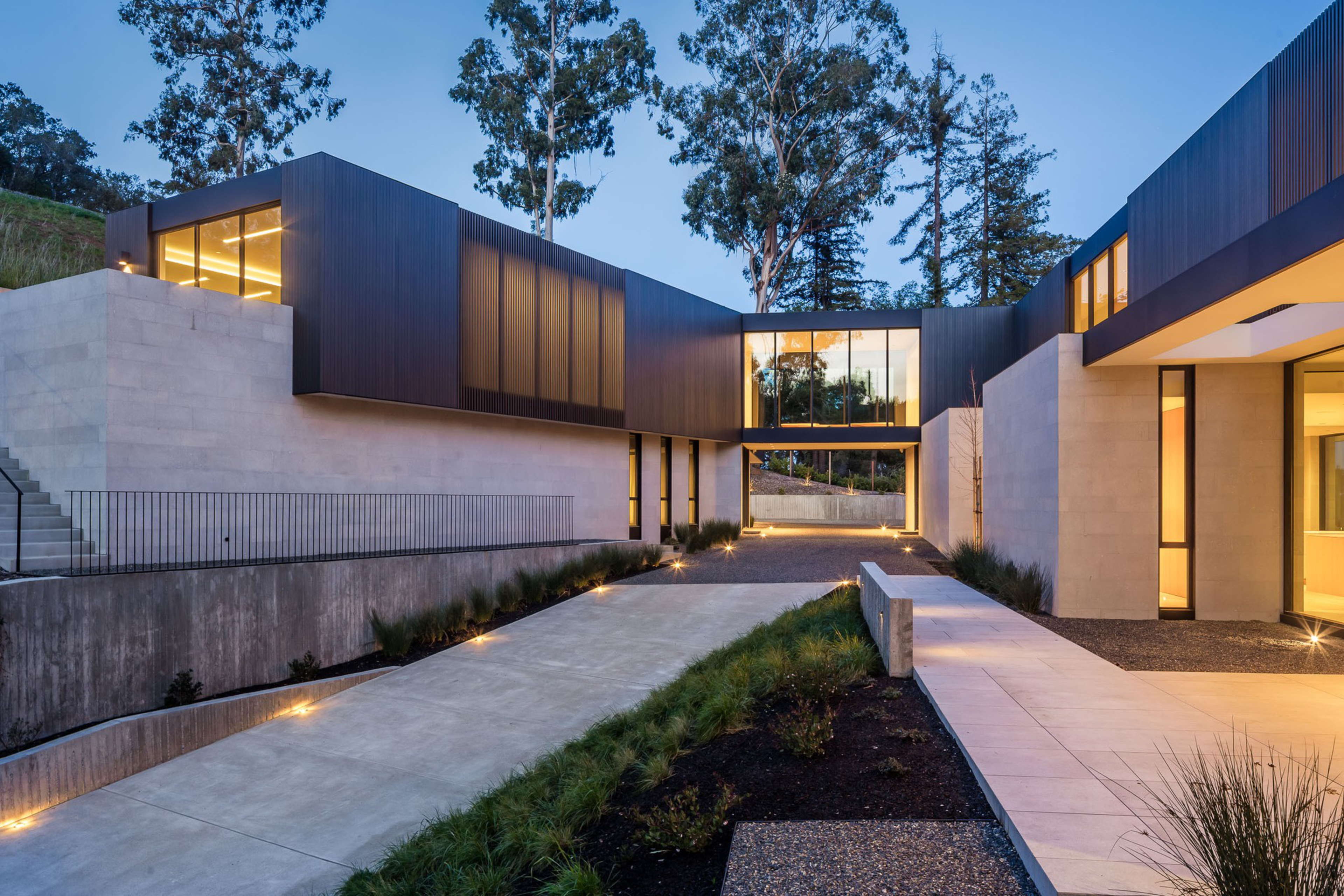 A modern house with angular design, concrete and dark panel walls, large windows, a driveway lined with grass, and outdoor lighting at dusk.