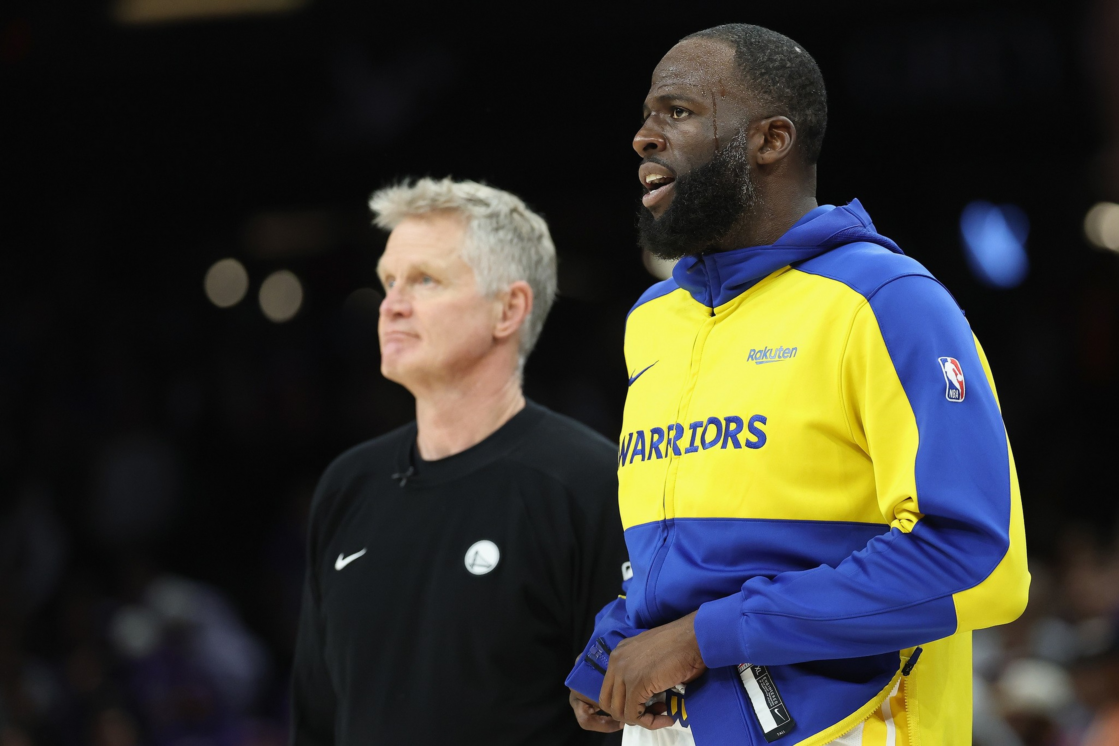 A man in a blue and yellow Warriors jacket stands next to a man in a black shirt with a white circular logo, both looking forward.