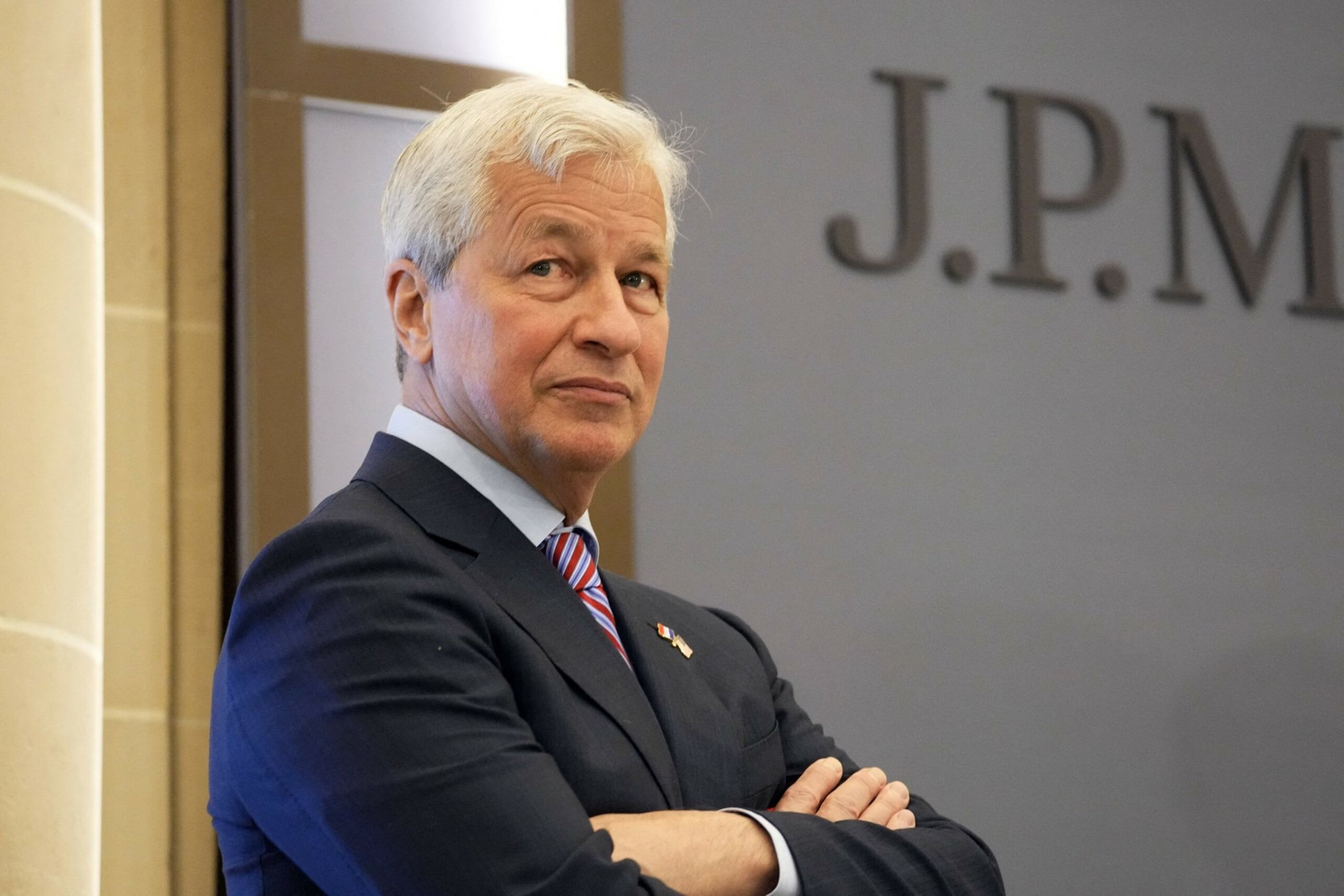 An older man in a suit with white hair is standing with arms crossed. The background shows part of a wall with "J.P. M" visible.