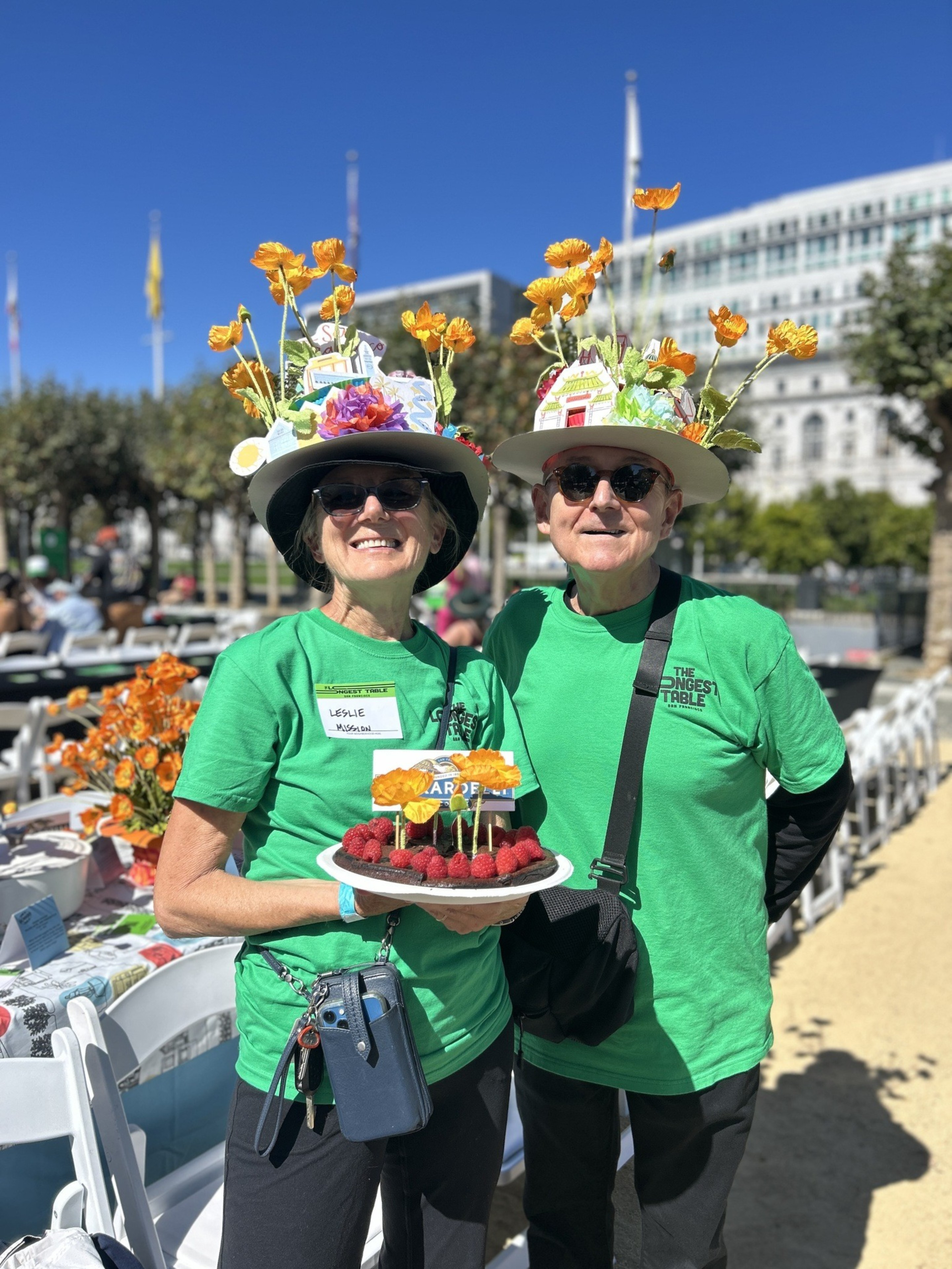 Two people wearing green shirts, sunglasses, and hats decorated with flowers smile while holding a plate of raspberries and orange flowers outdoors.