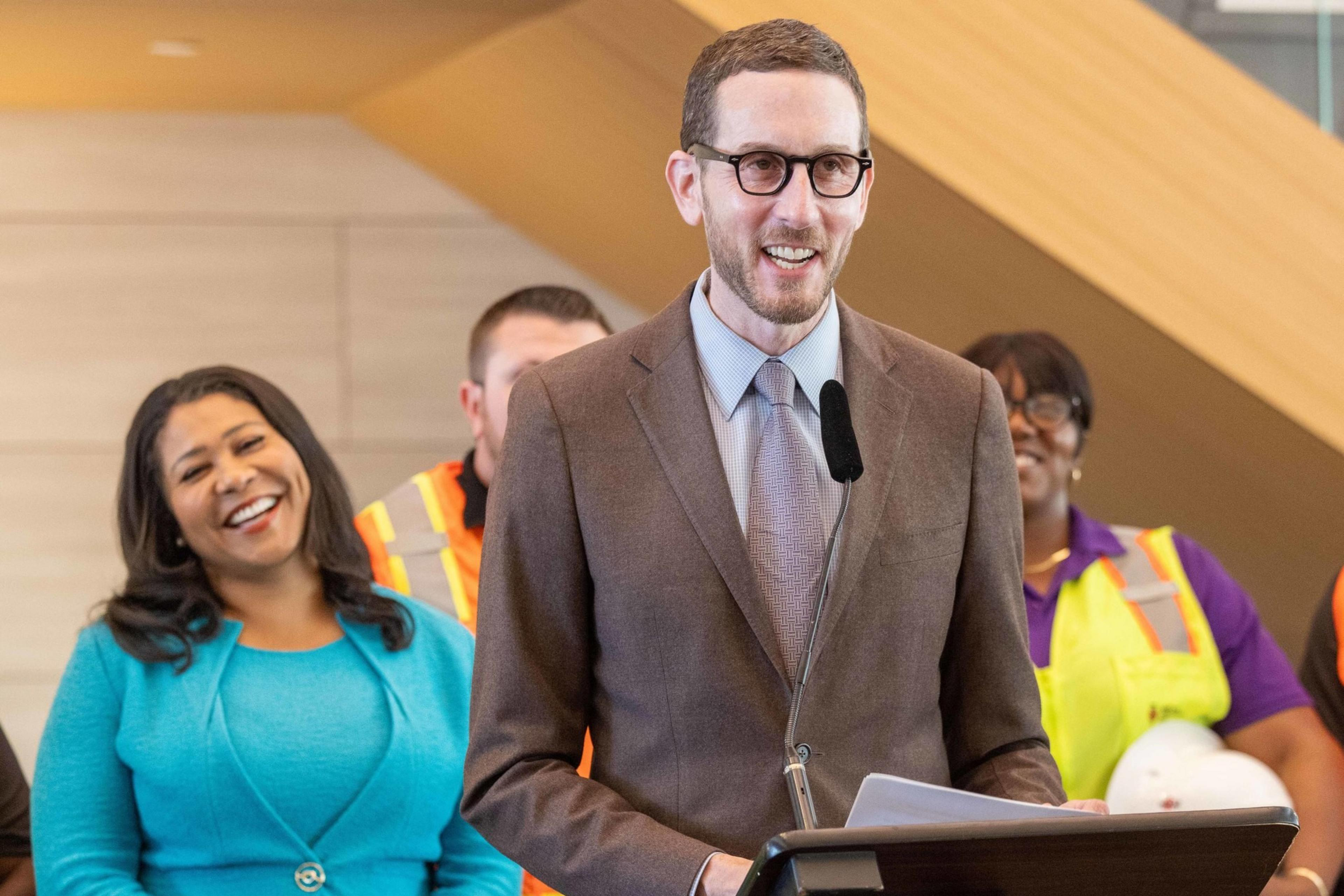 A man in a brown suit speaks at a podium with a microphone, while a woman in a blue outfit smiles behind him. Several people in construction attire stand in the background.
