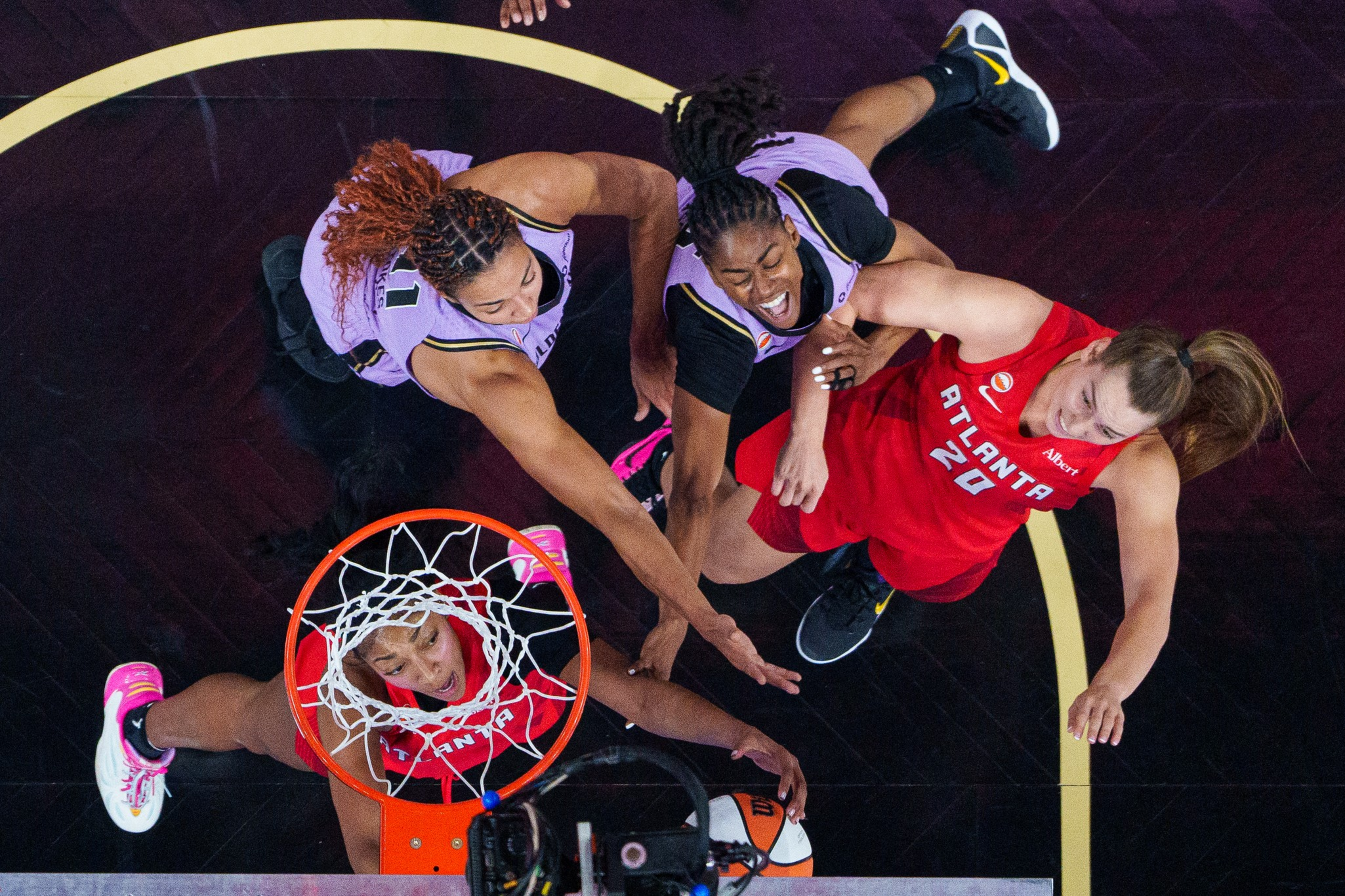 Two players in purple uniforms and two in red uniforms compete for a basketball near the hoop during a game, with the ball close to the rim.