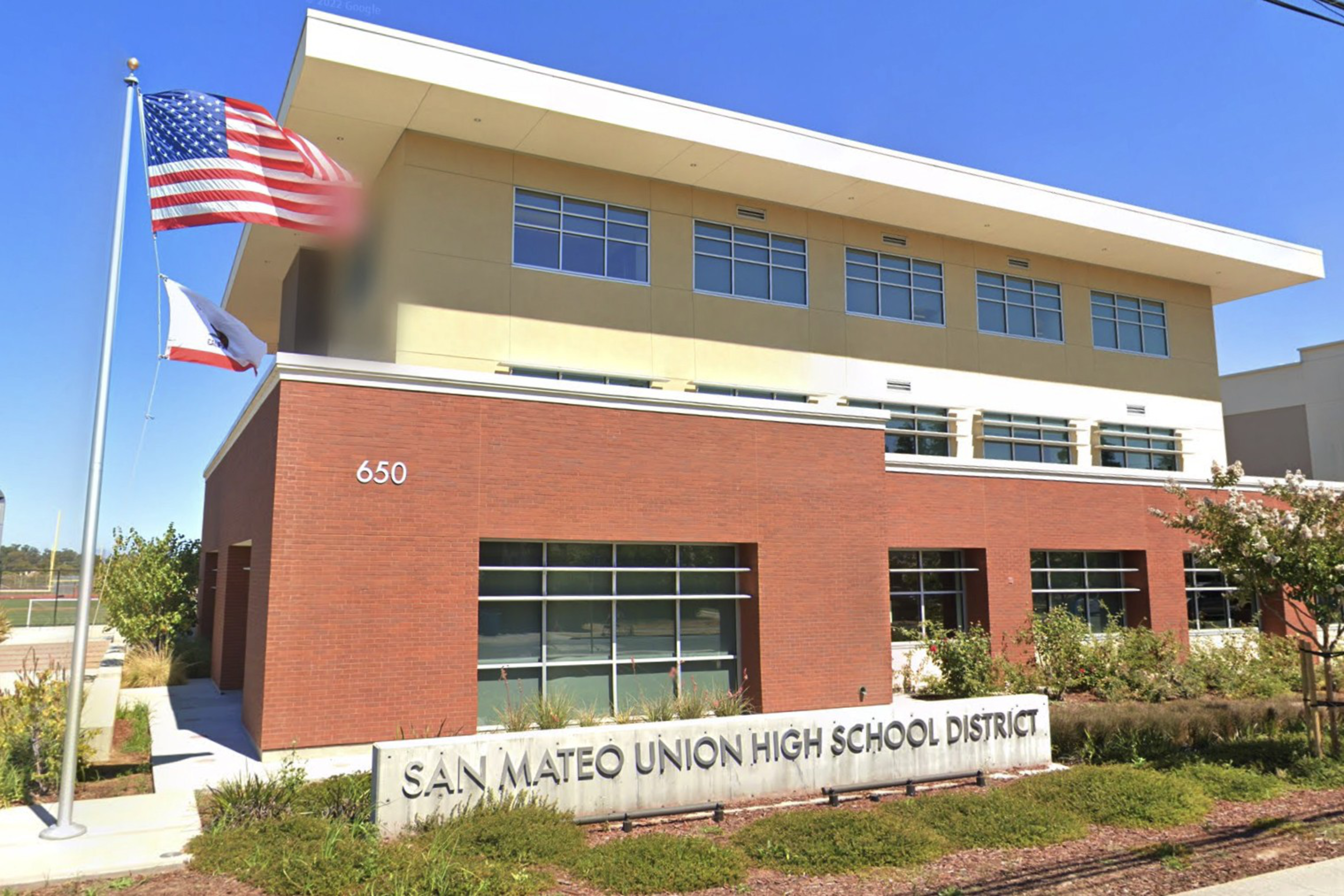 A modern two-story building with red brick and beige walls, windows, and a sign reading "San Mateo Union High School District" with American and California flags nearby.