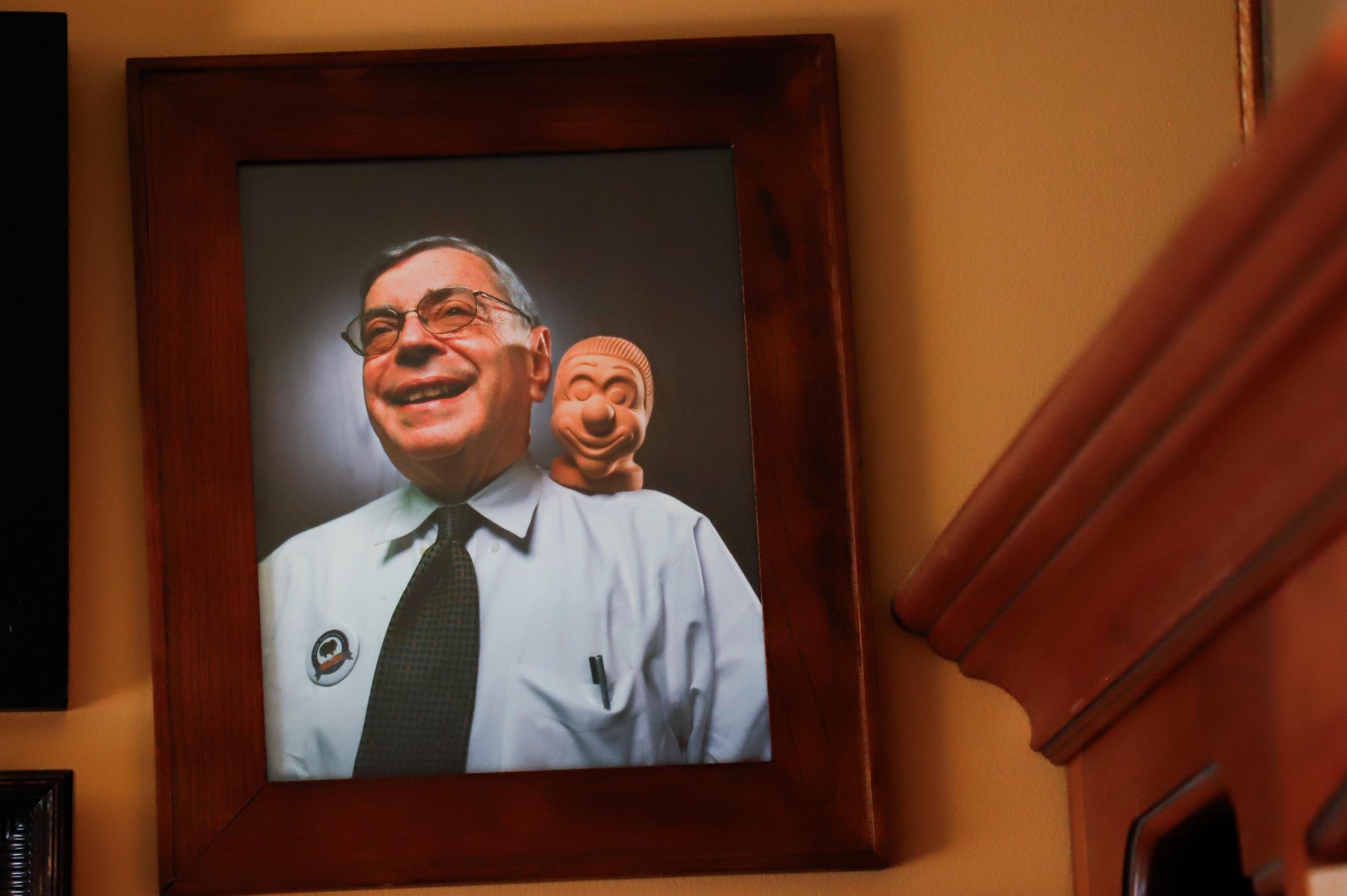 A framed photo of a smiling older man in glasses and a tie has a cartoonish clay figure peeking over his shoulder, both facing forward.