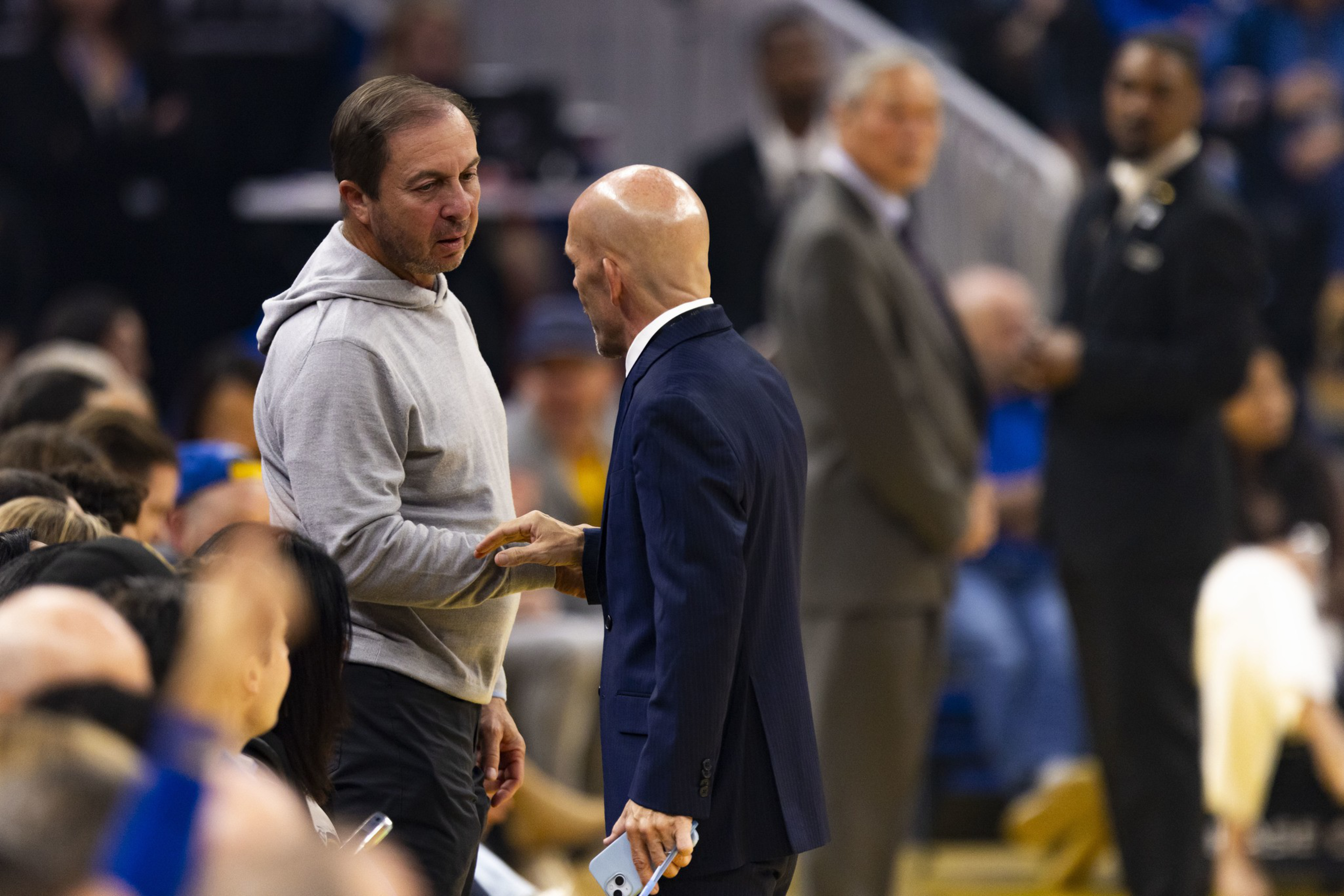 A man in a gray hoodie shakes hands with a bald man in a dark suit holding a phone during a crowded indoor event.