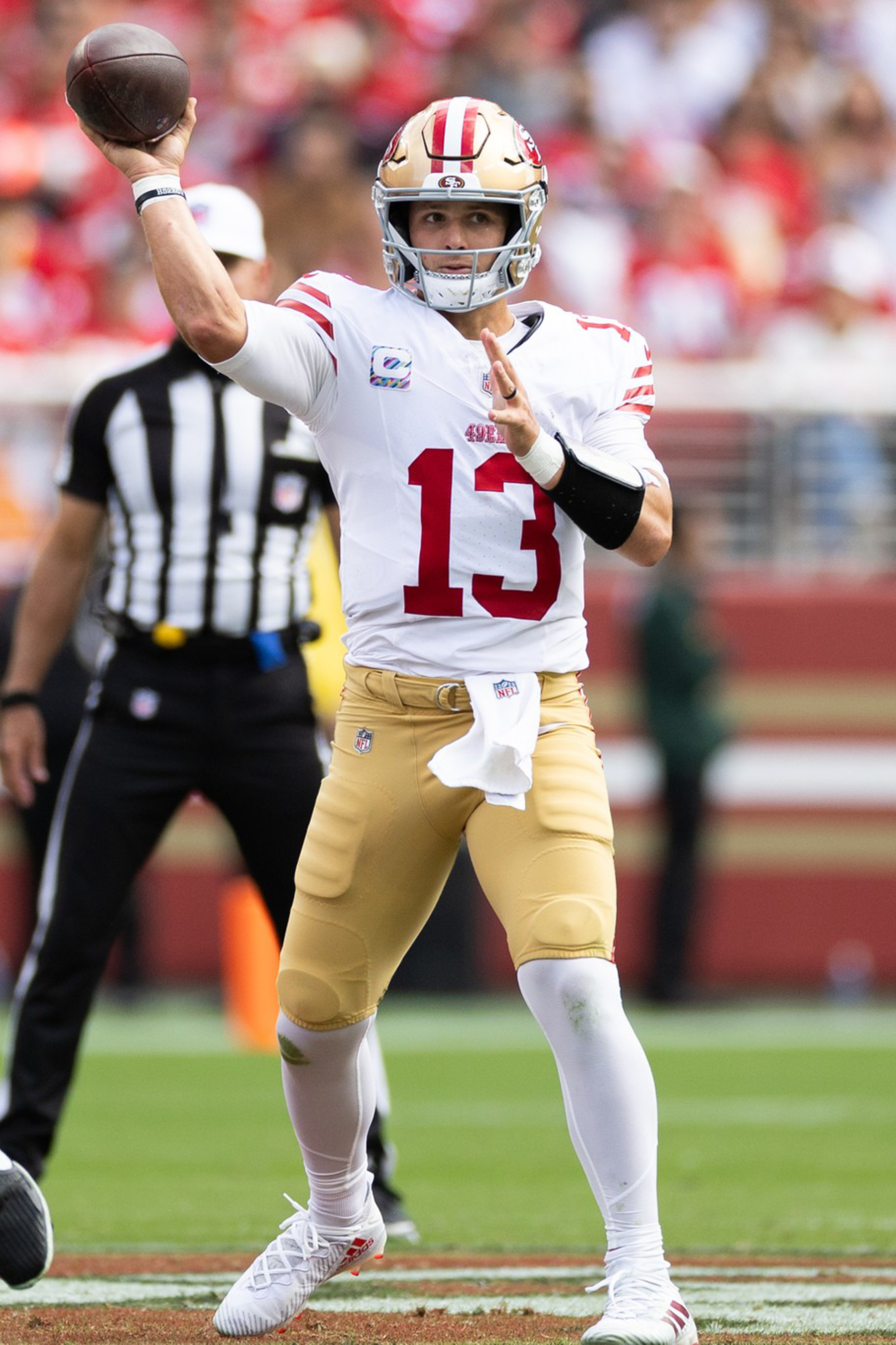 A football player wearing a number 13 jersey prepares to throw a pass during a game, with a teammate blocking and a referee watching behind him.