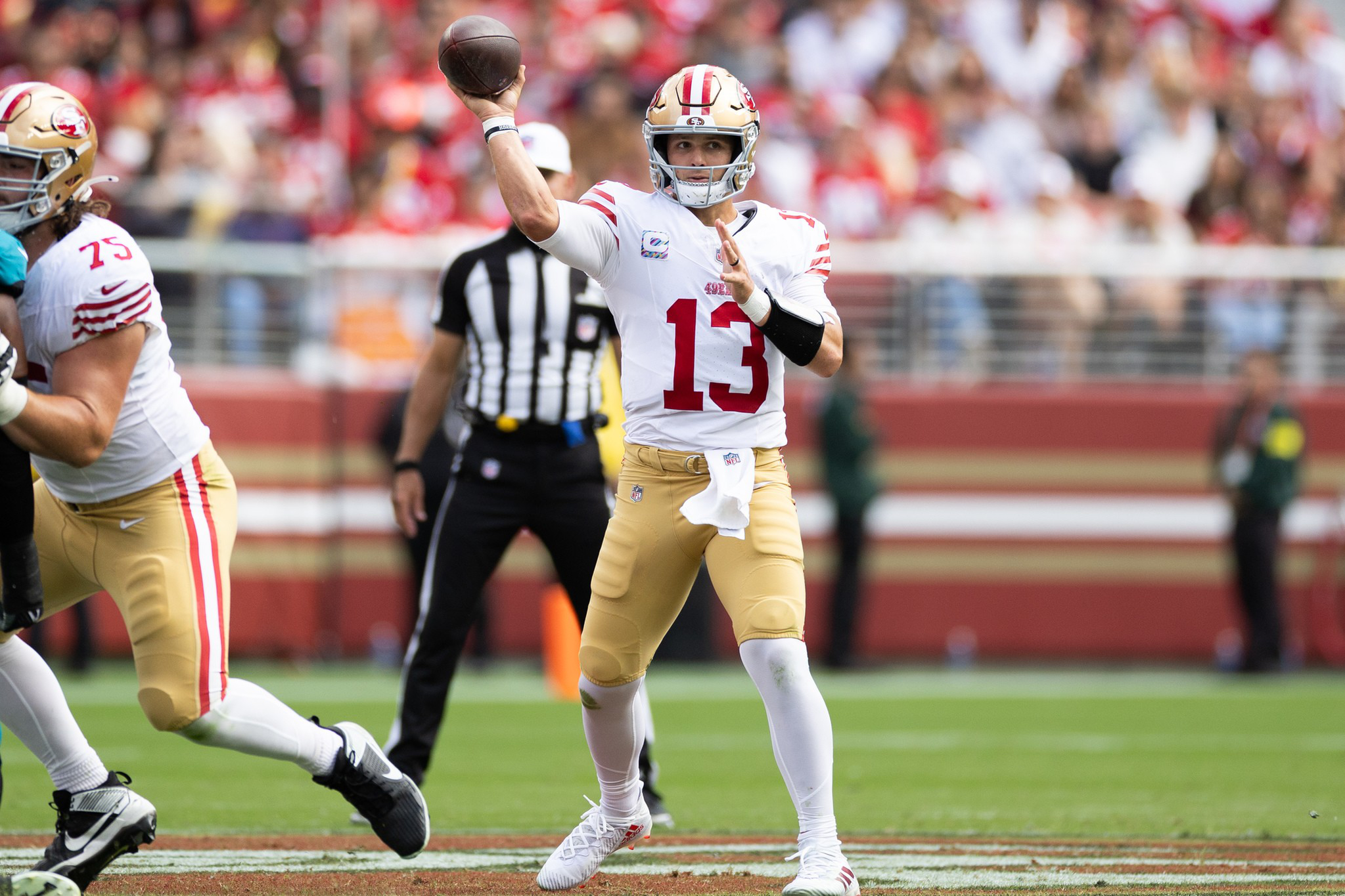 A football player wearing a number 13 jersey prepares to throw a pass during a game, with a teammate blocking and a referee watching behind him.