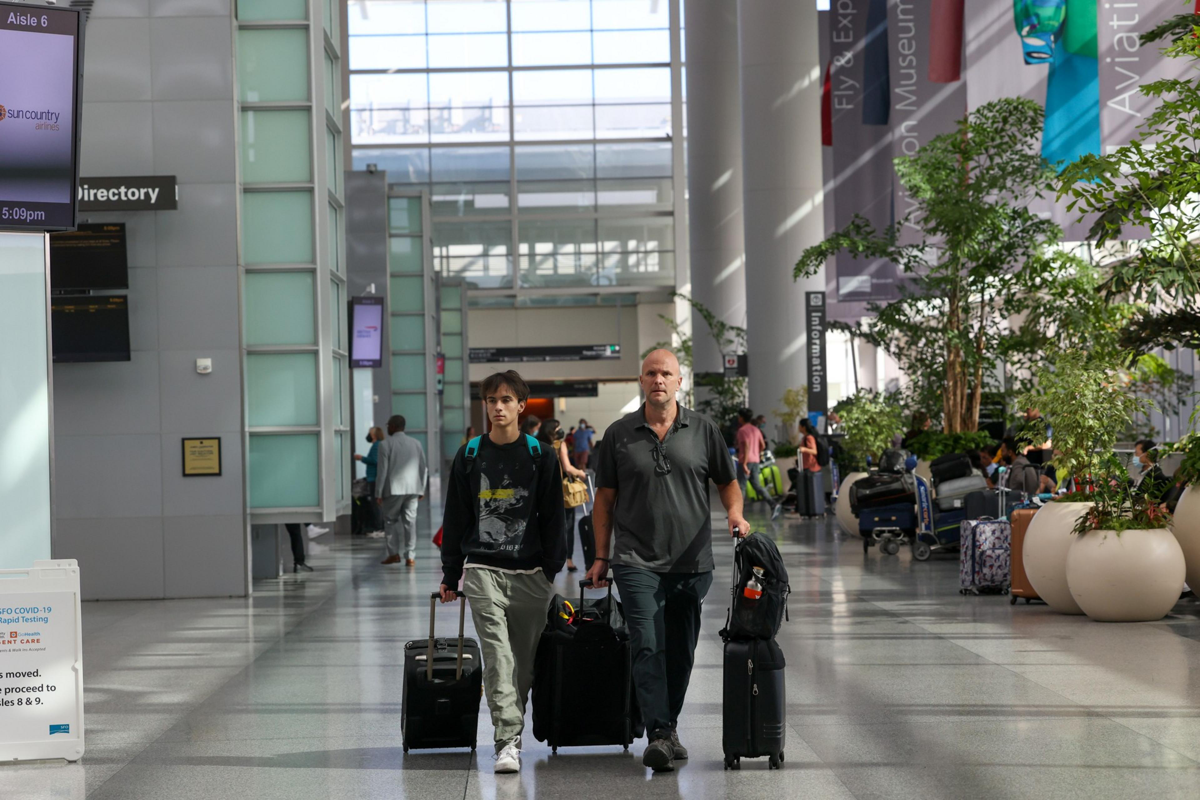 Two men walk through a bright airport terminal, each pulling a rolling suitcase, with plants and other travelers visible in the background.
