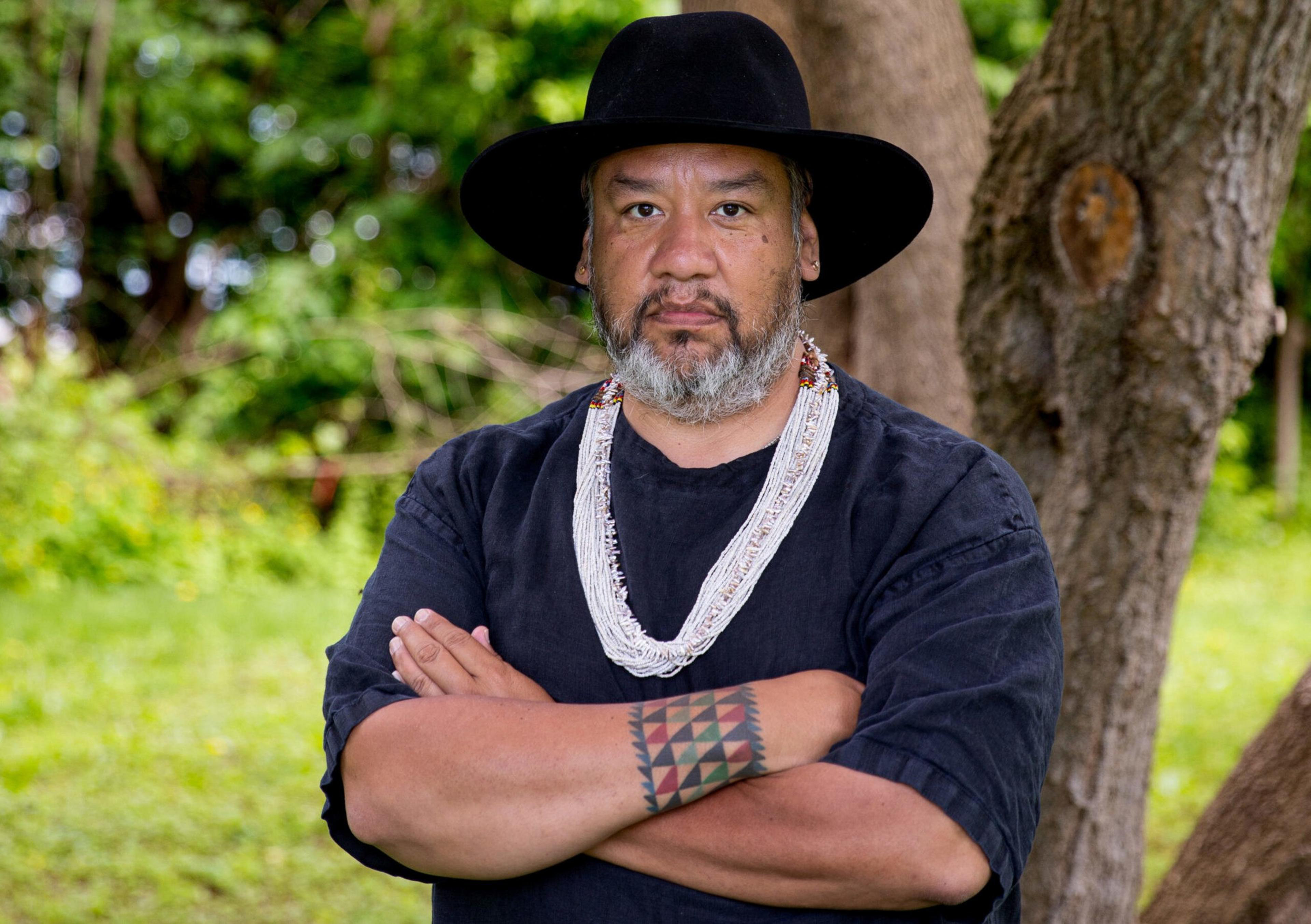 A man with a black hat and beard stands with arms crossed, wearing a black shirt, white beaded necklace, and a colorful geometric tattoo on his forearm.