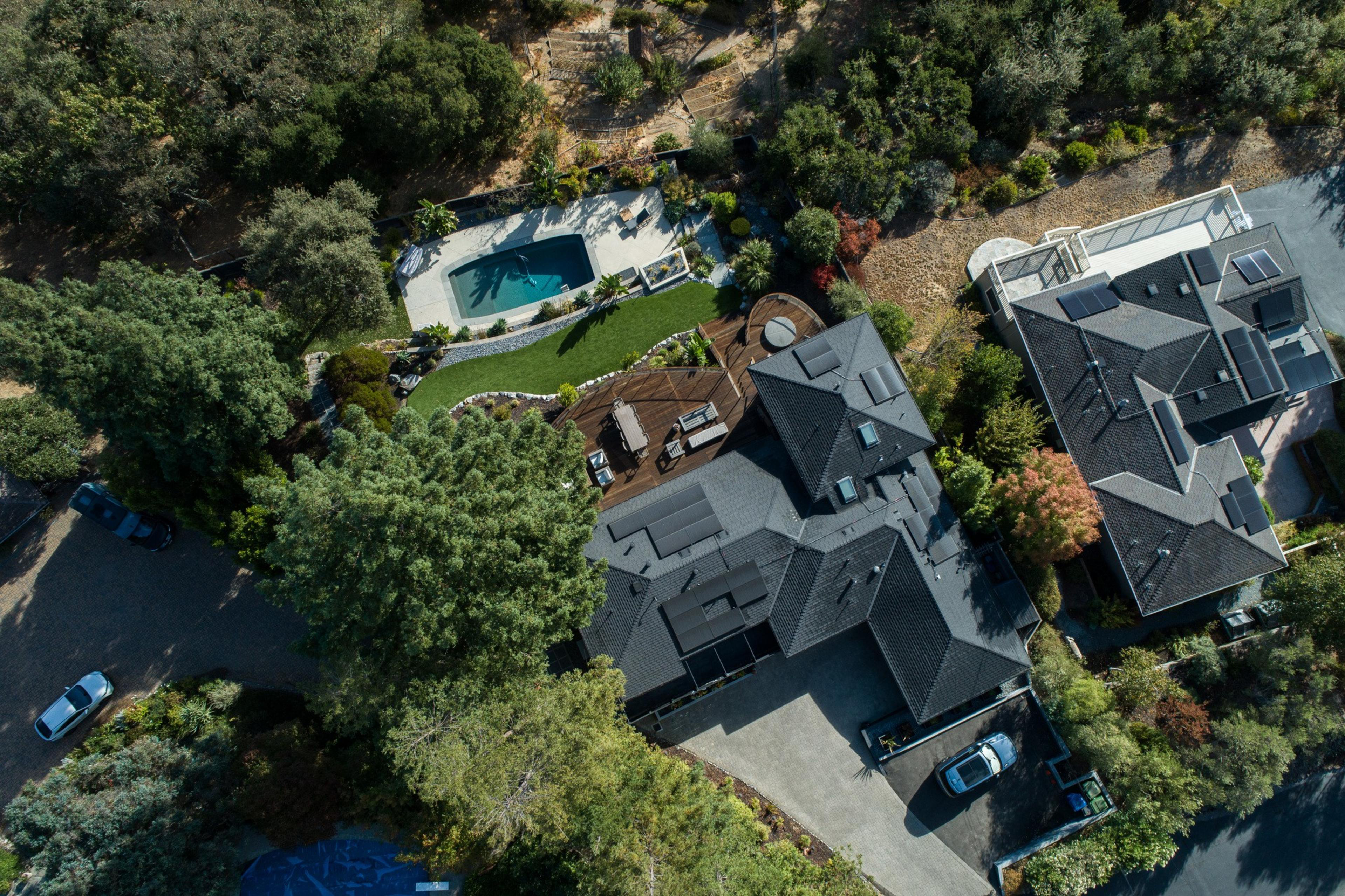 The image shows an aerial view of a house with a dark roof, surrounded by a patio, a green lawn, a swimming pool, and trees, with neighboring homes nearby.