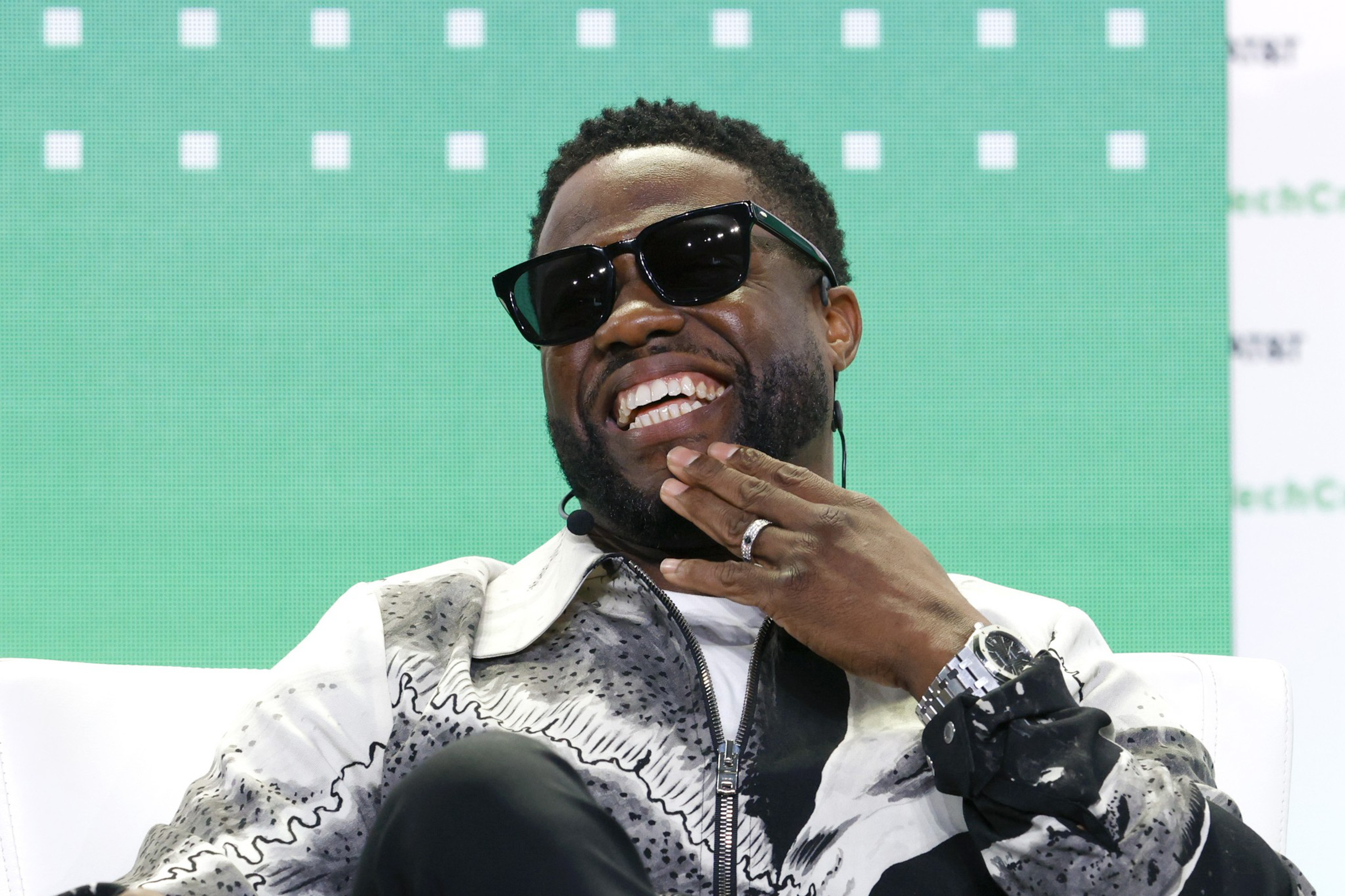 A man with a beard wearing black sunglasses and a patterned jacket smiles broadly while resting his hand near his face.