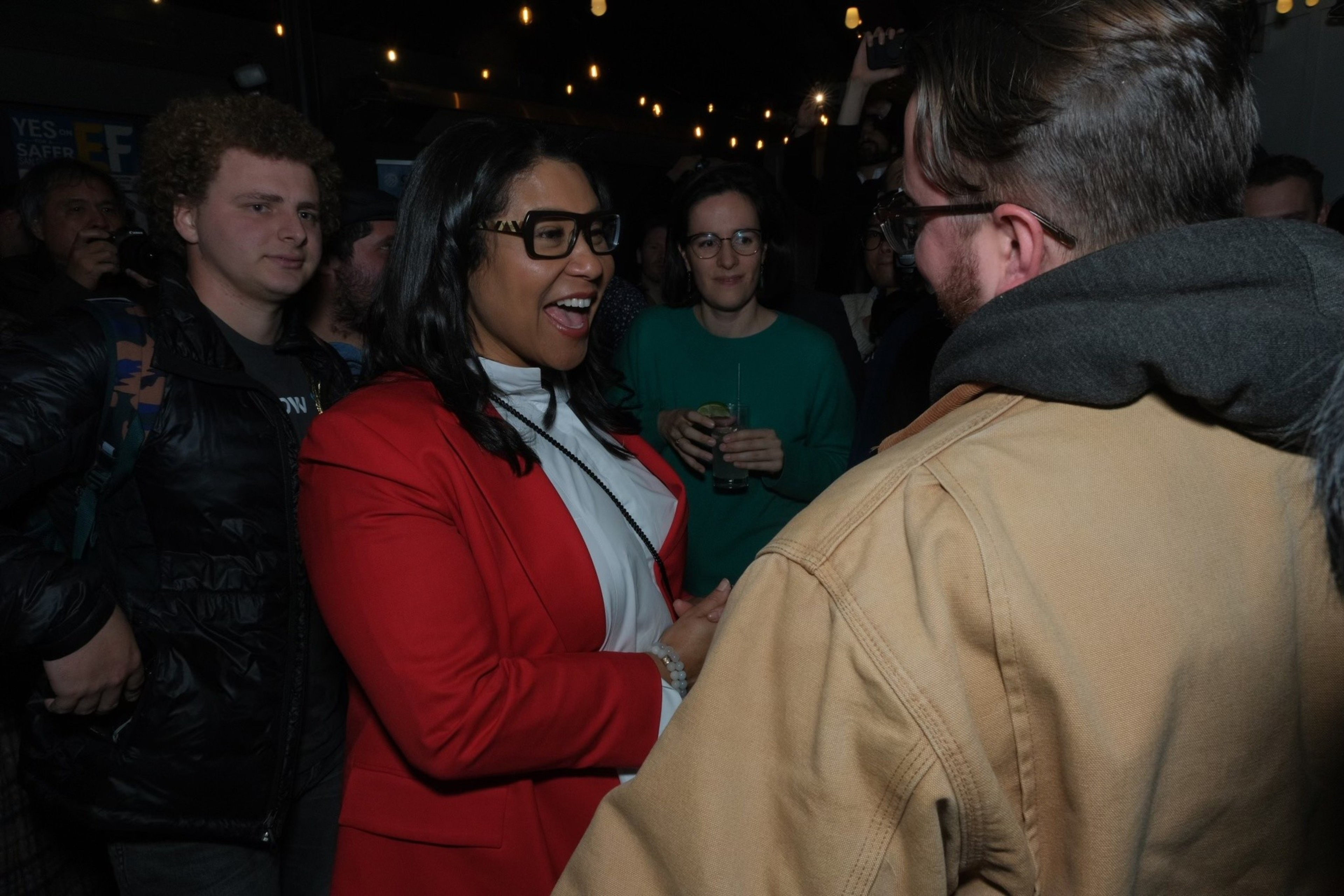 A woman in a red blazer is smiling and shaking hands with someone in a beige jacket at a crowded indoor event.
