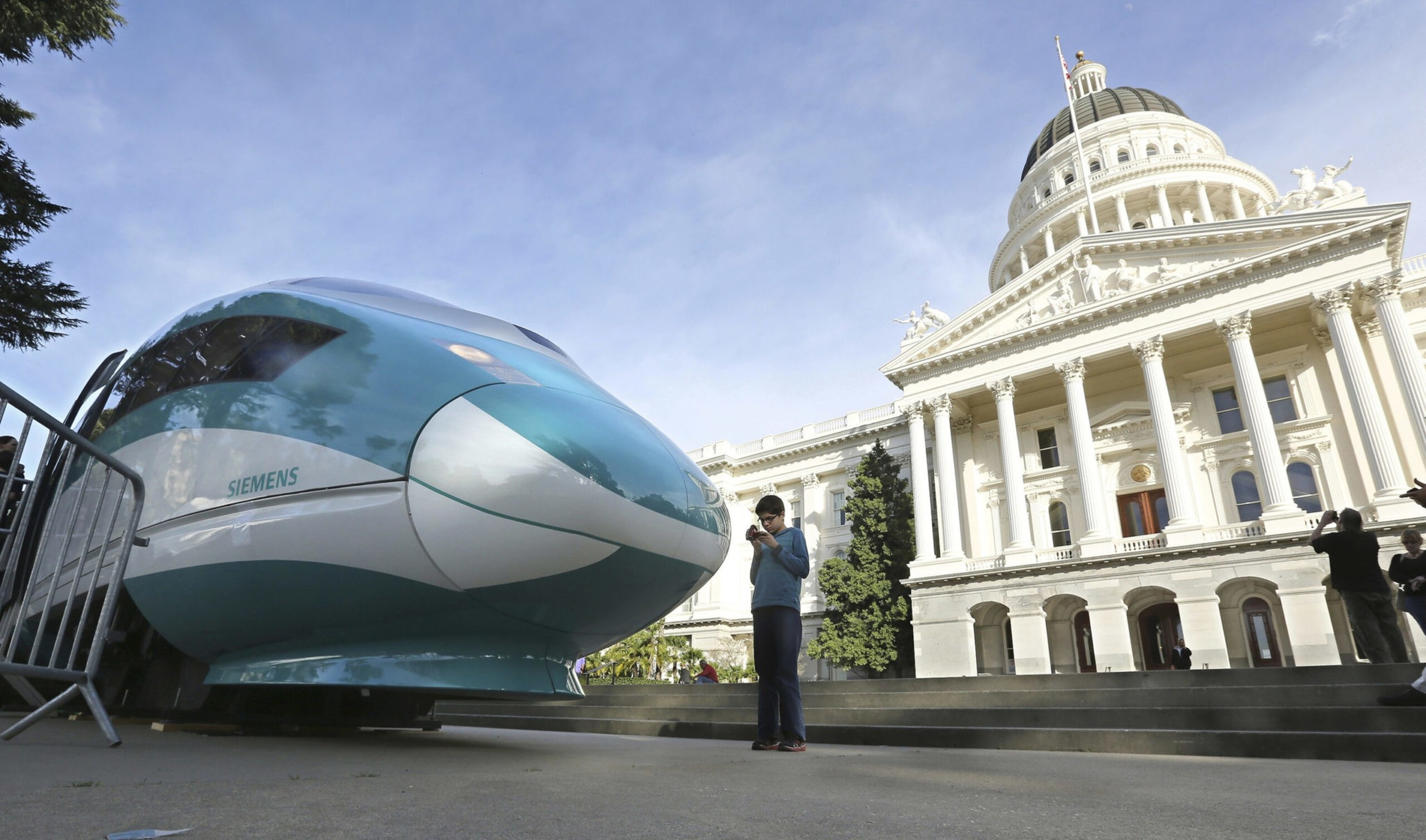 A sleek Siemens train nose is displayed outdoors near a large white domed government building, with people taking photos nearby.