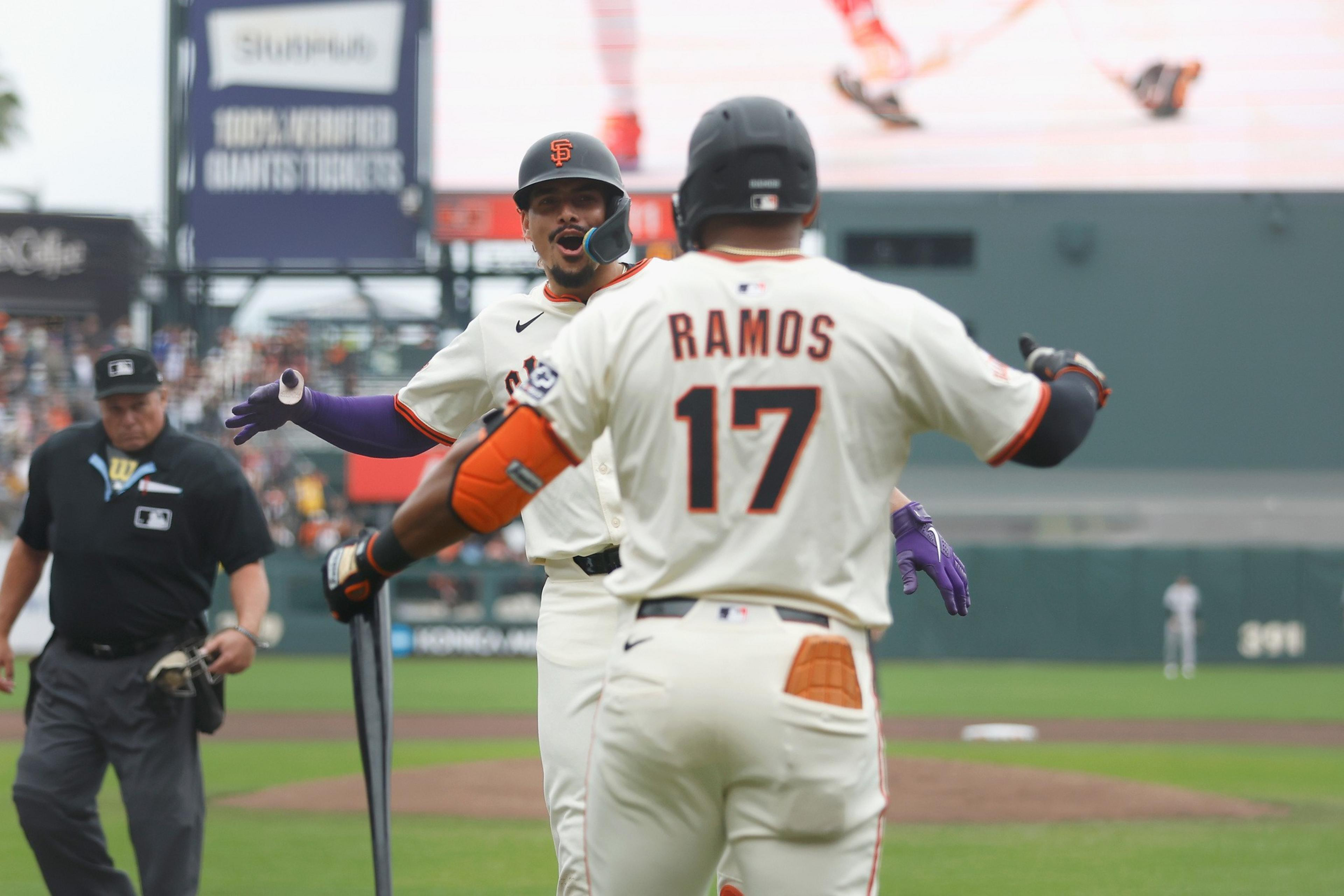 Two San Francisco Giants players celebrate on the field; one holds a bat with an umpire walking nearby.