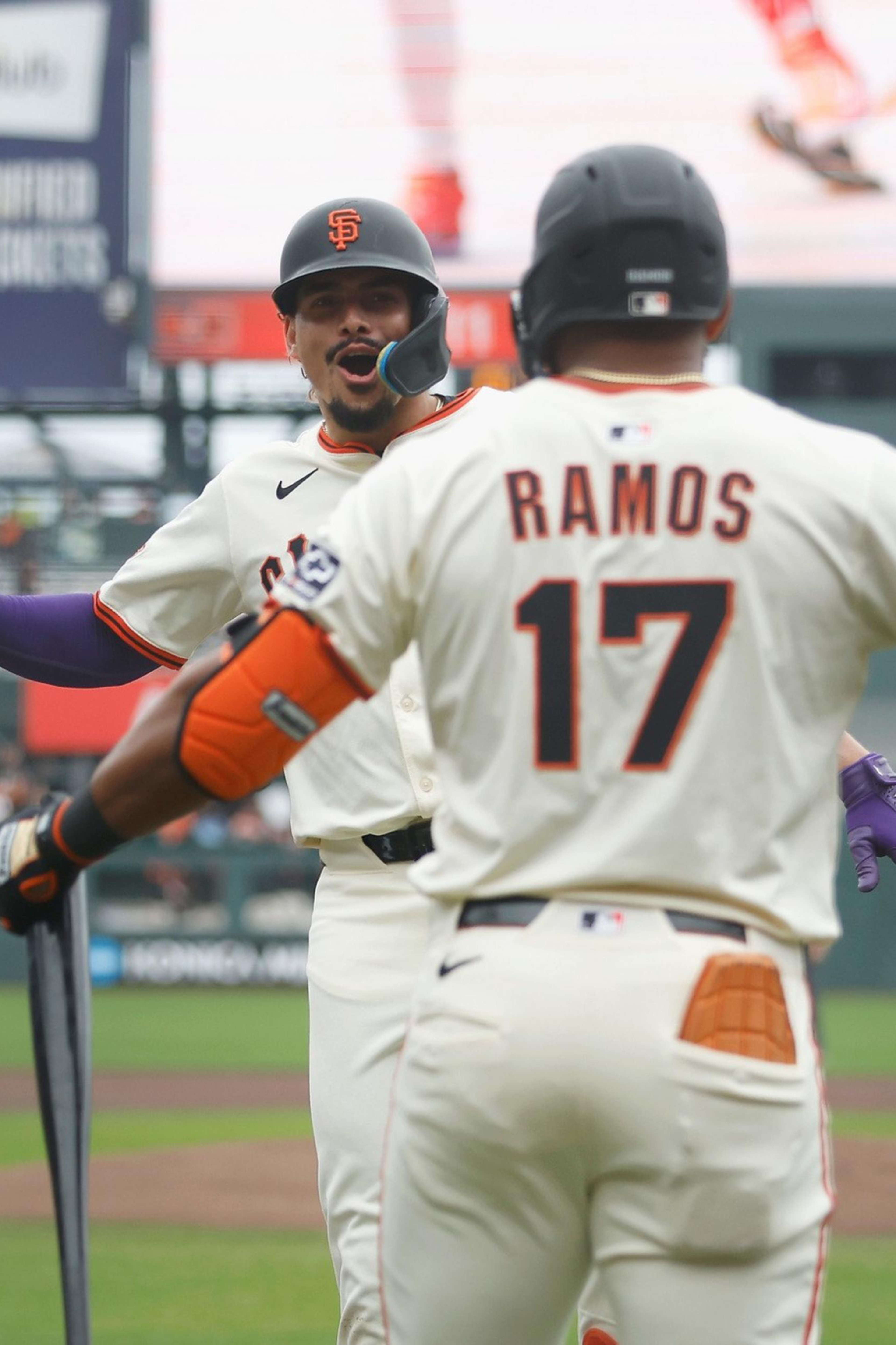Two San Francisco Giants players celebrate on the field; one holds a bat with an umpire walking nearby.