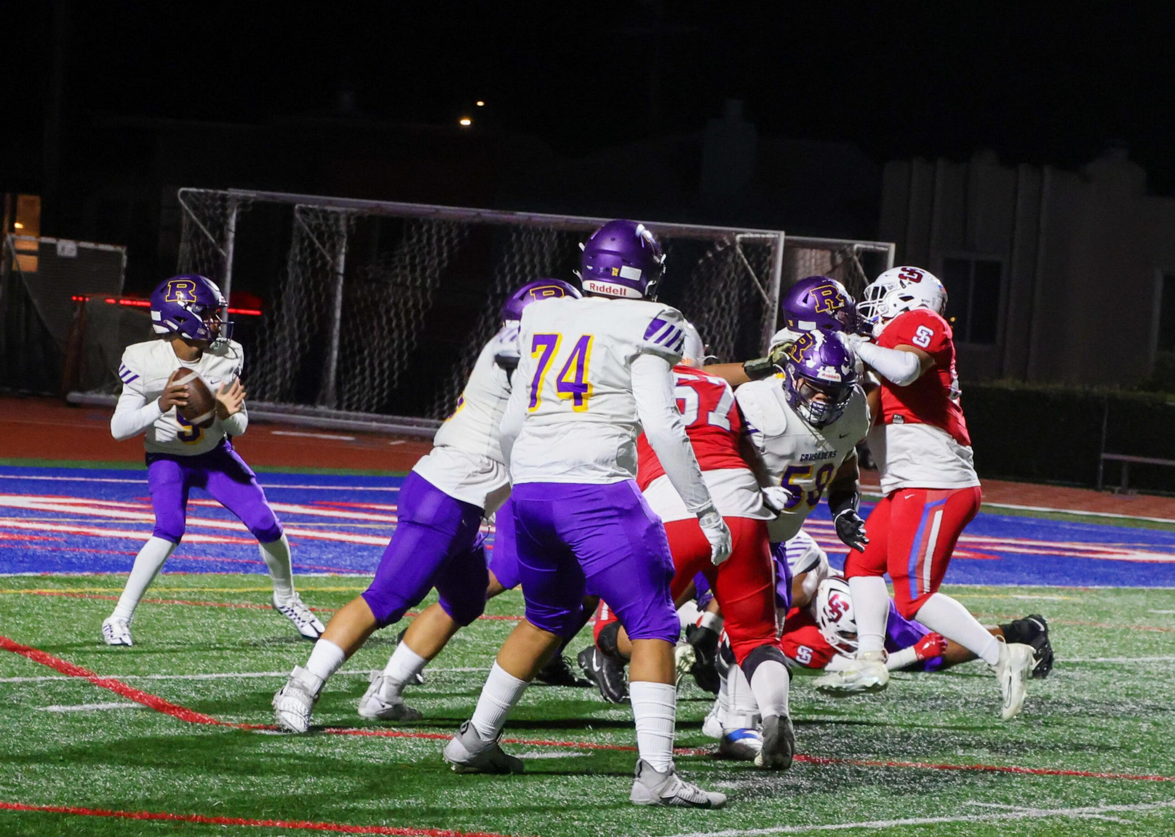 A football quarterback in white and purple prepares to throw while teammates block defenders in red on a brightly lit field at night.