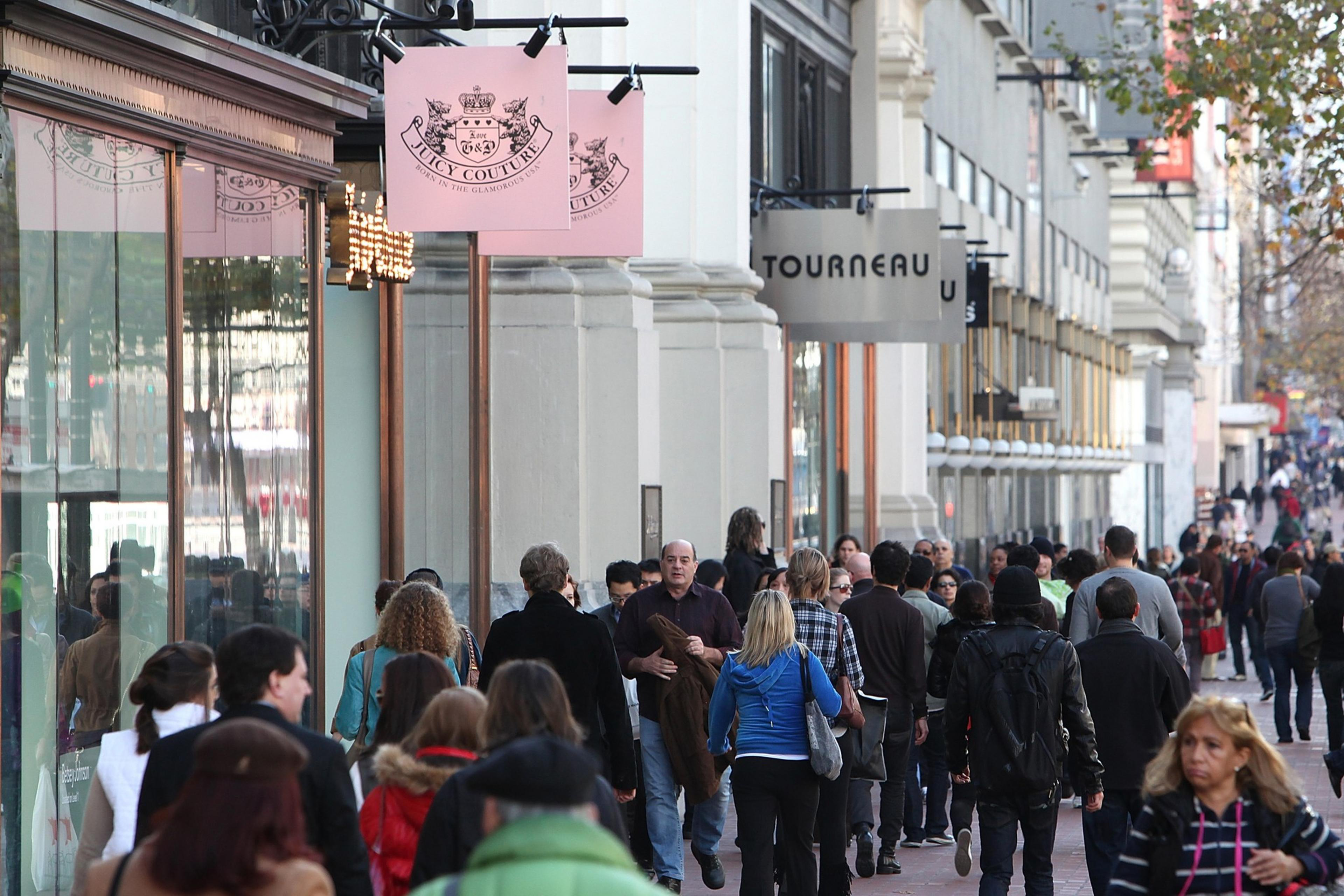 A busy urban sidewalk filled with diverse people walking past storefronts with signs, like "Juicy Couture," on a clear day.