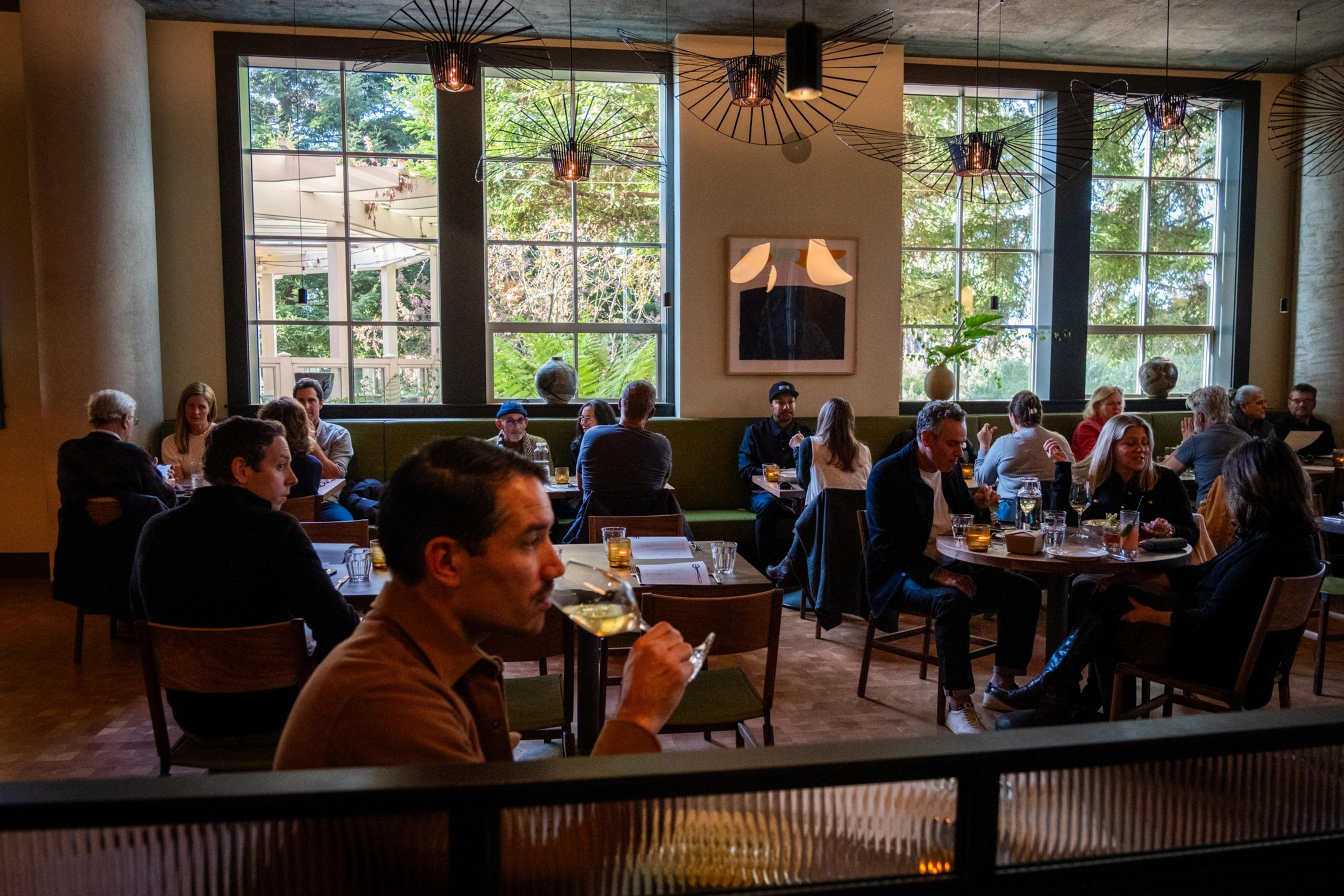 A bustling restaurant with large windows and modern lighting, filled with diners enjoying meals at tables. Green plants and warm lighting add a cozy ambiance.
