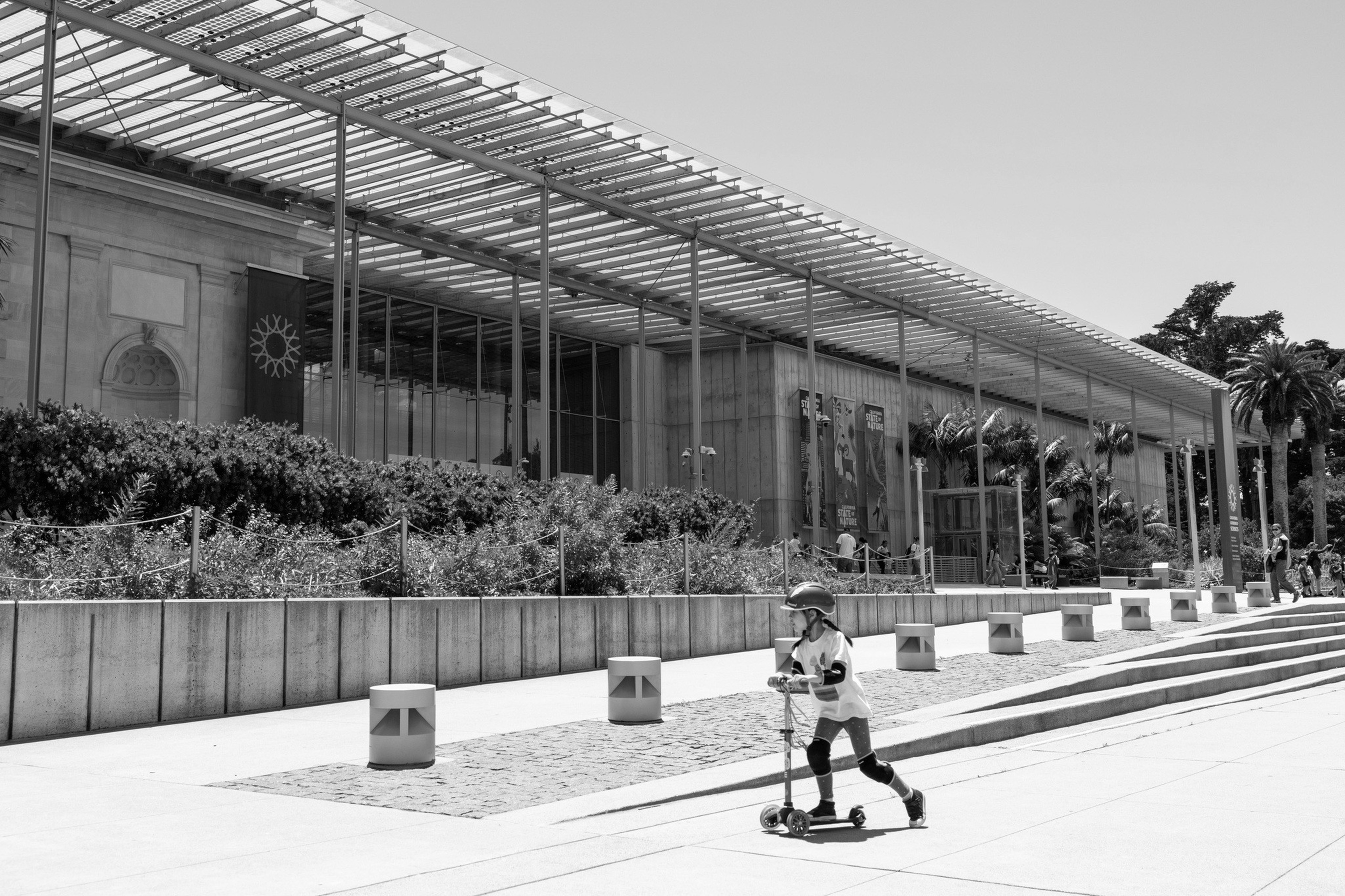 A child wearing a helmet rides a scooter along a wide sidewalk in front of a large building with glass walls and vertical poles.
