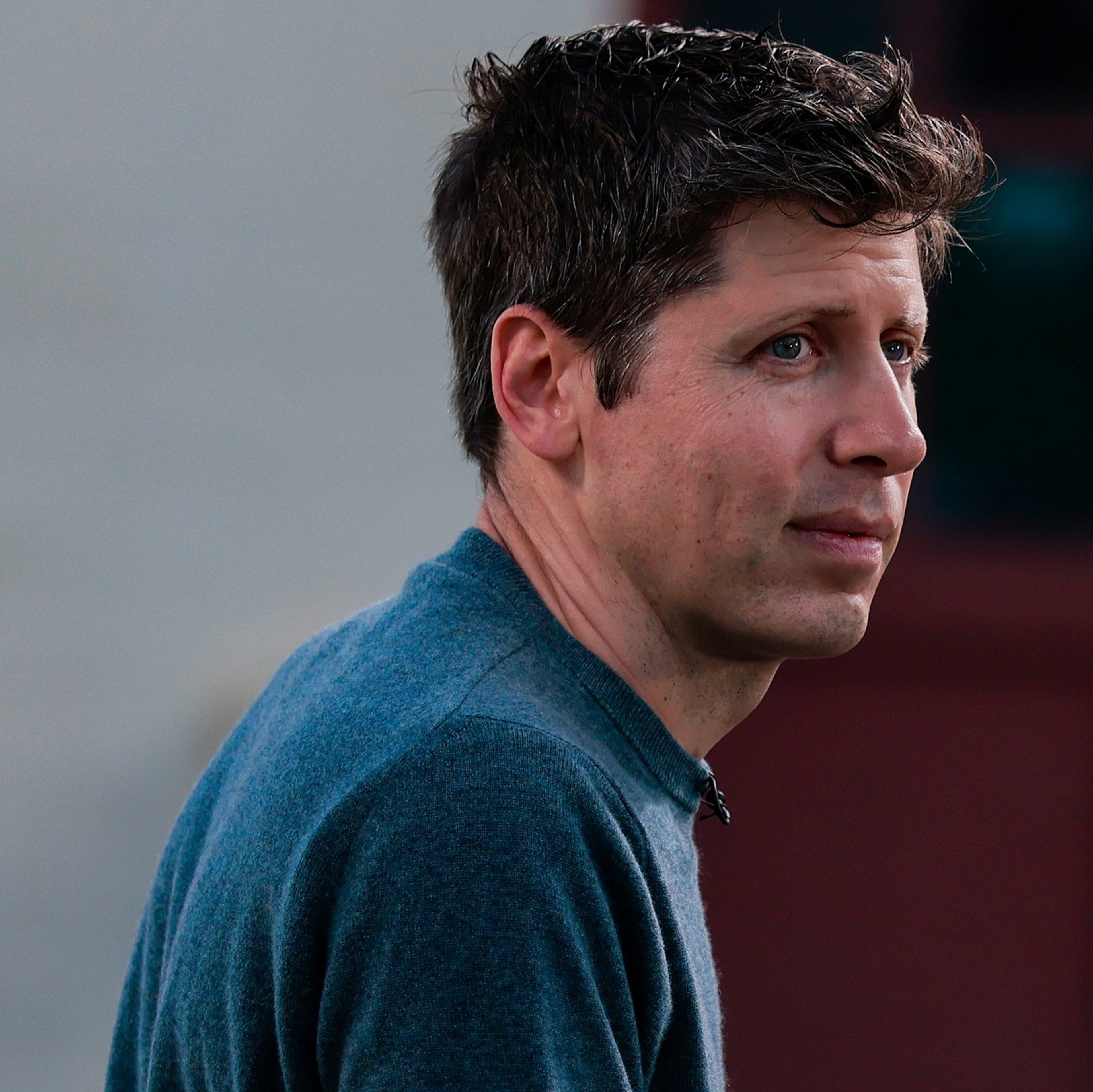 A man with short, curly brown hair wears a blue sweater, looking thoughtfully to his right against a blurred background.