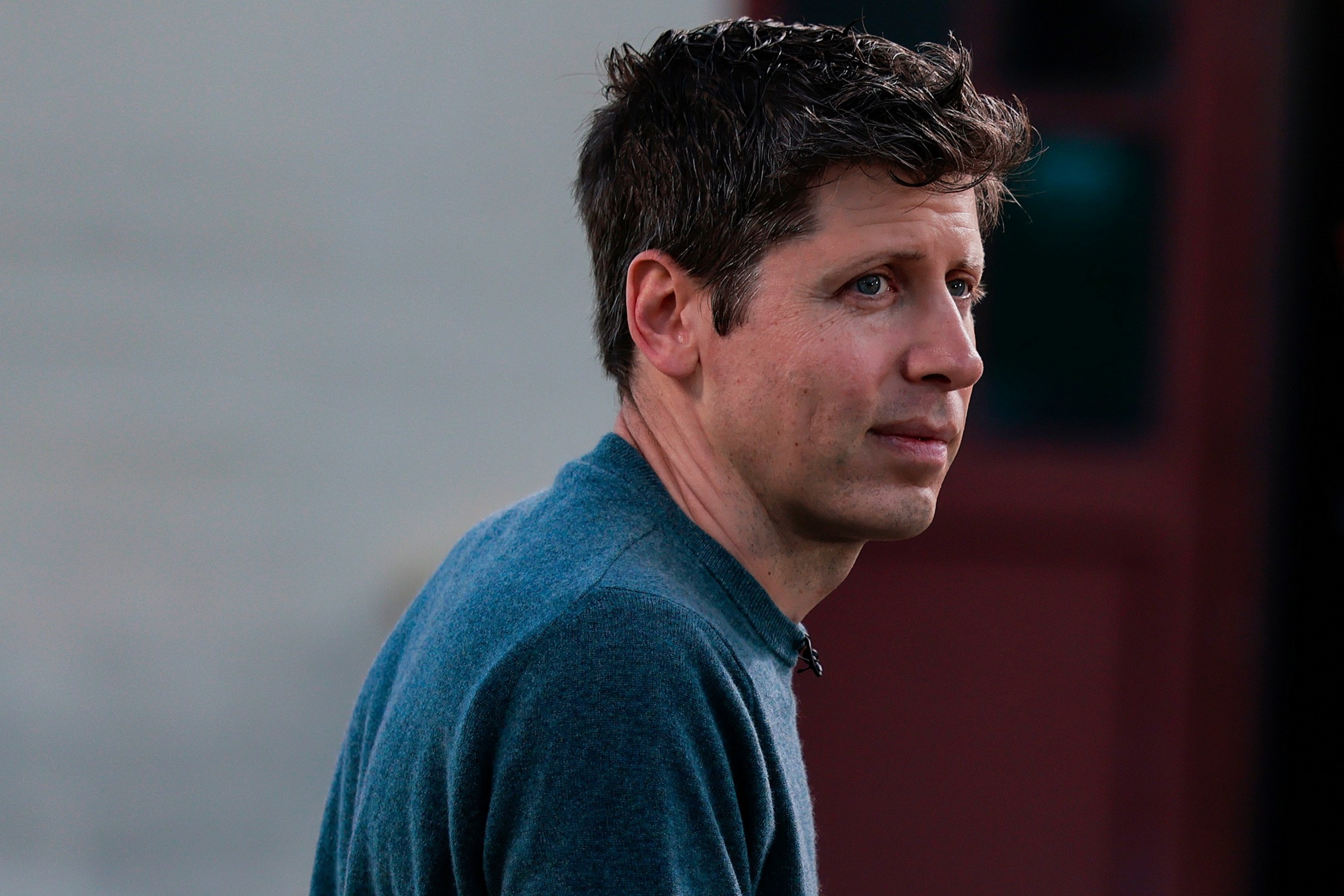 A man with short, curly brown hair wears a blue sweater, looking thoughtfully to his right against a blurred background.