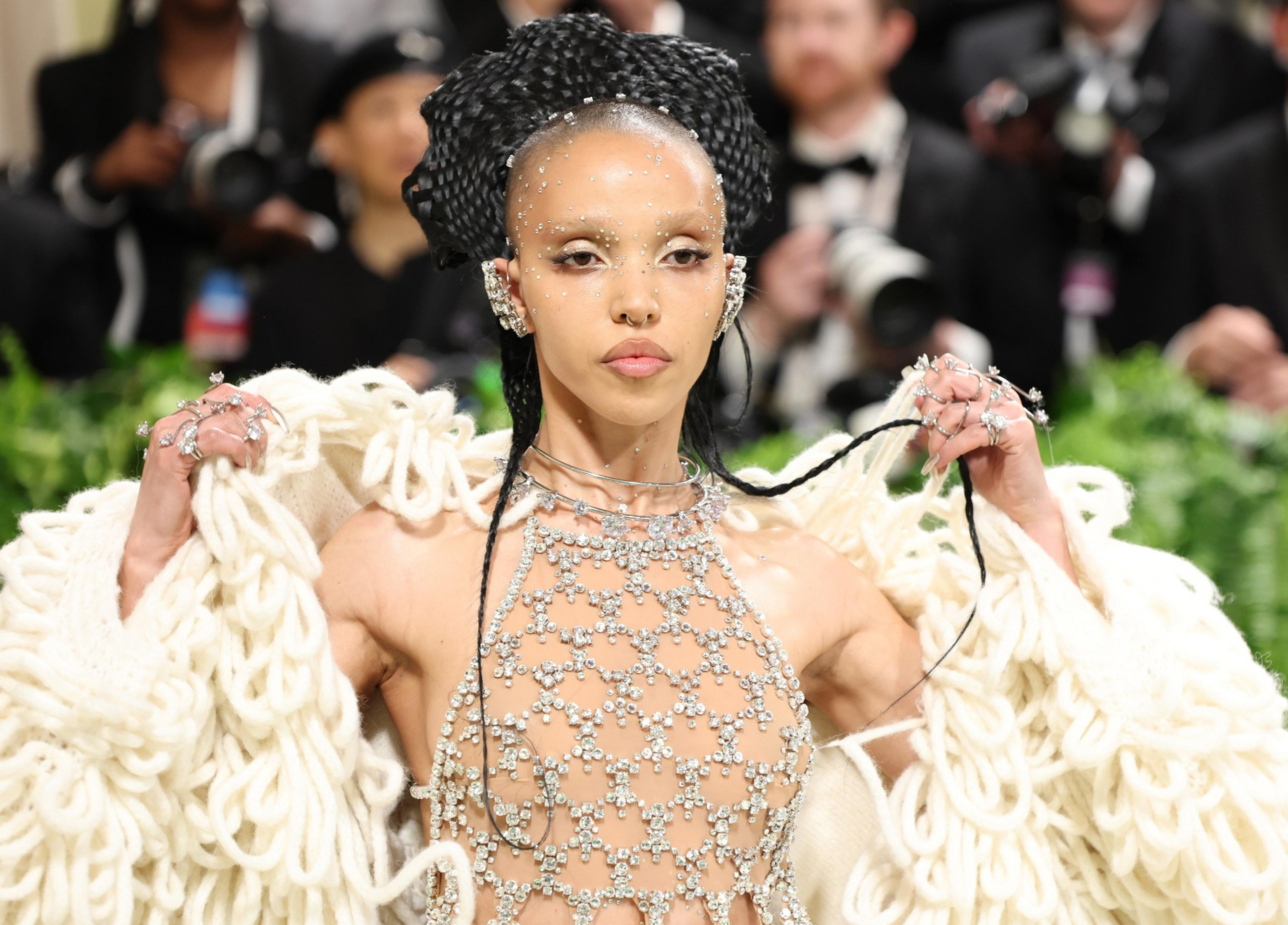 A person wears a sparkling, gem-studded outfit with elaborate jewelry and a dramatic, braided hairstyle. Photographers are visible in the background.