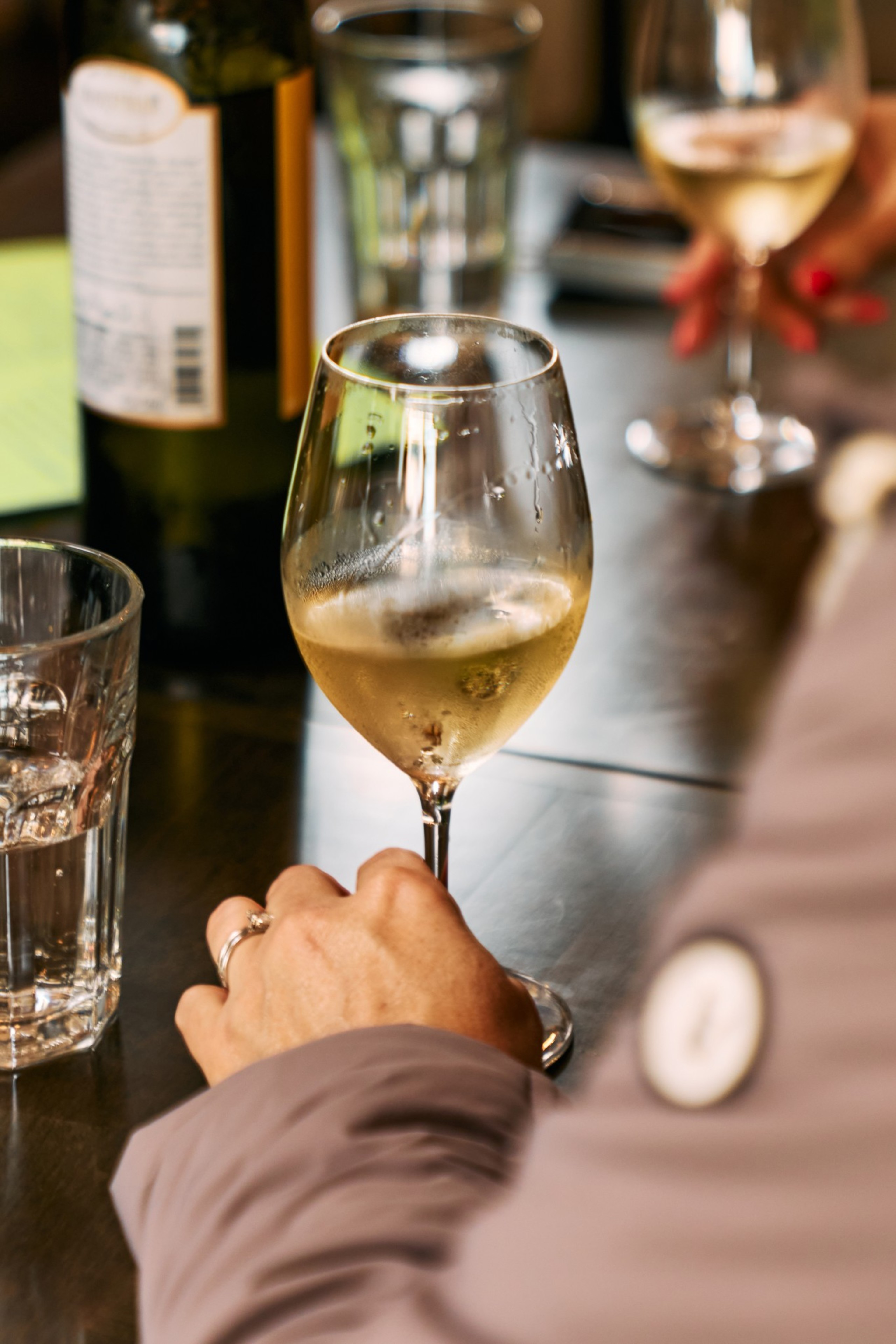 A hand wearing a ring holds a glass of white wine on a table with a water glass and a wine bottle nearby. Another person with red nails holds a similar glass.