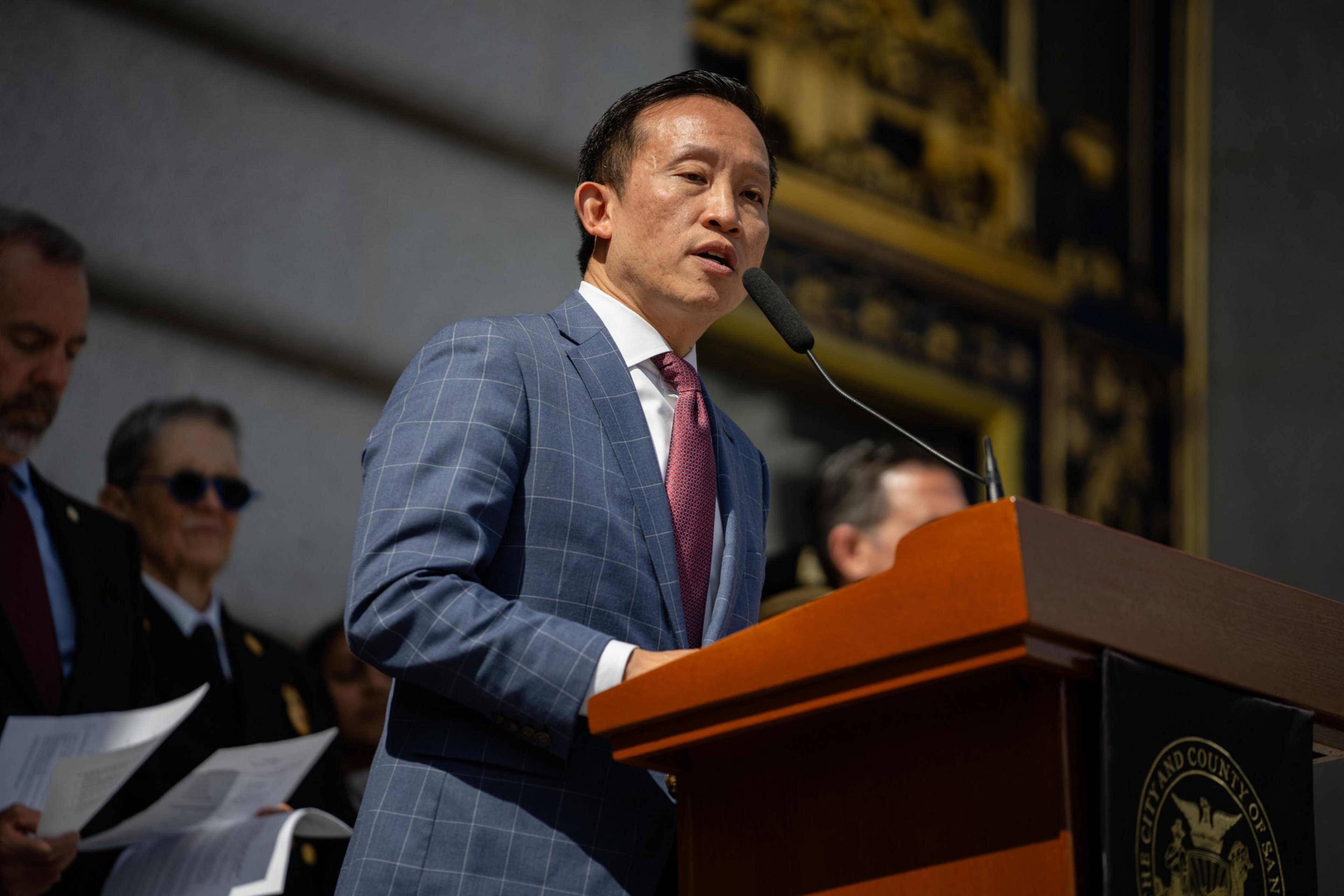 A man in a checkered suit speaks at a podium with people and a seal behind him.