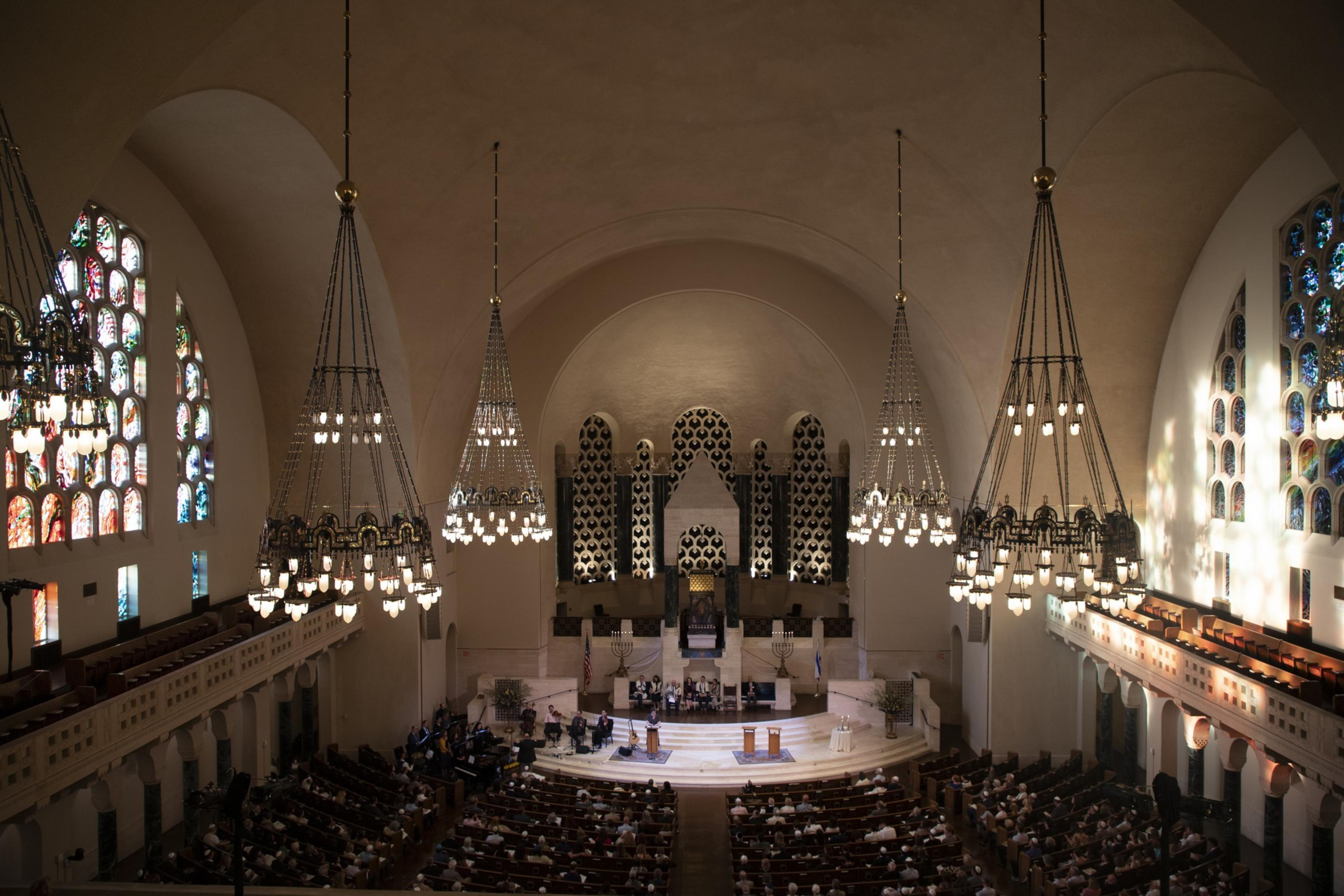A large synagogue with stained glass windows, grand chandeliers, and a stage. People are seated in pews, attending a service at the front where a choir is performing.