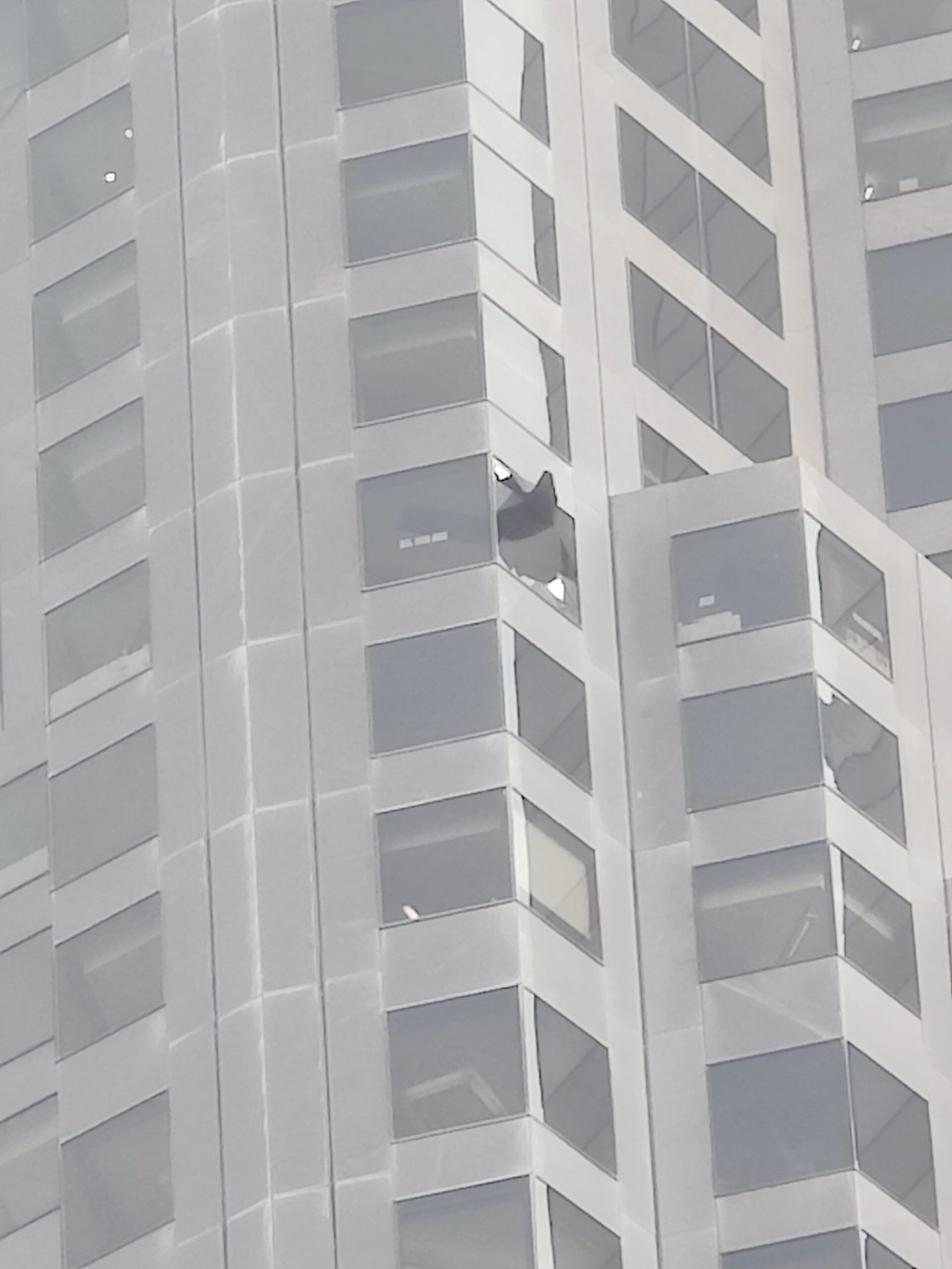 A high-rise building has one window on an upper floor that is broken, with glass shards missing and some still hanging.