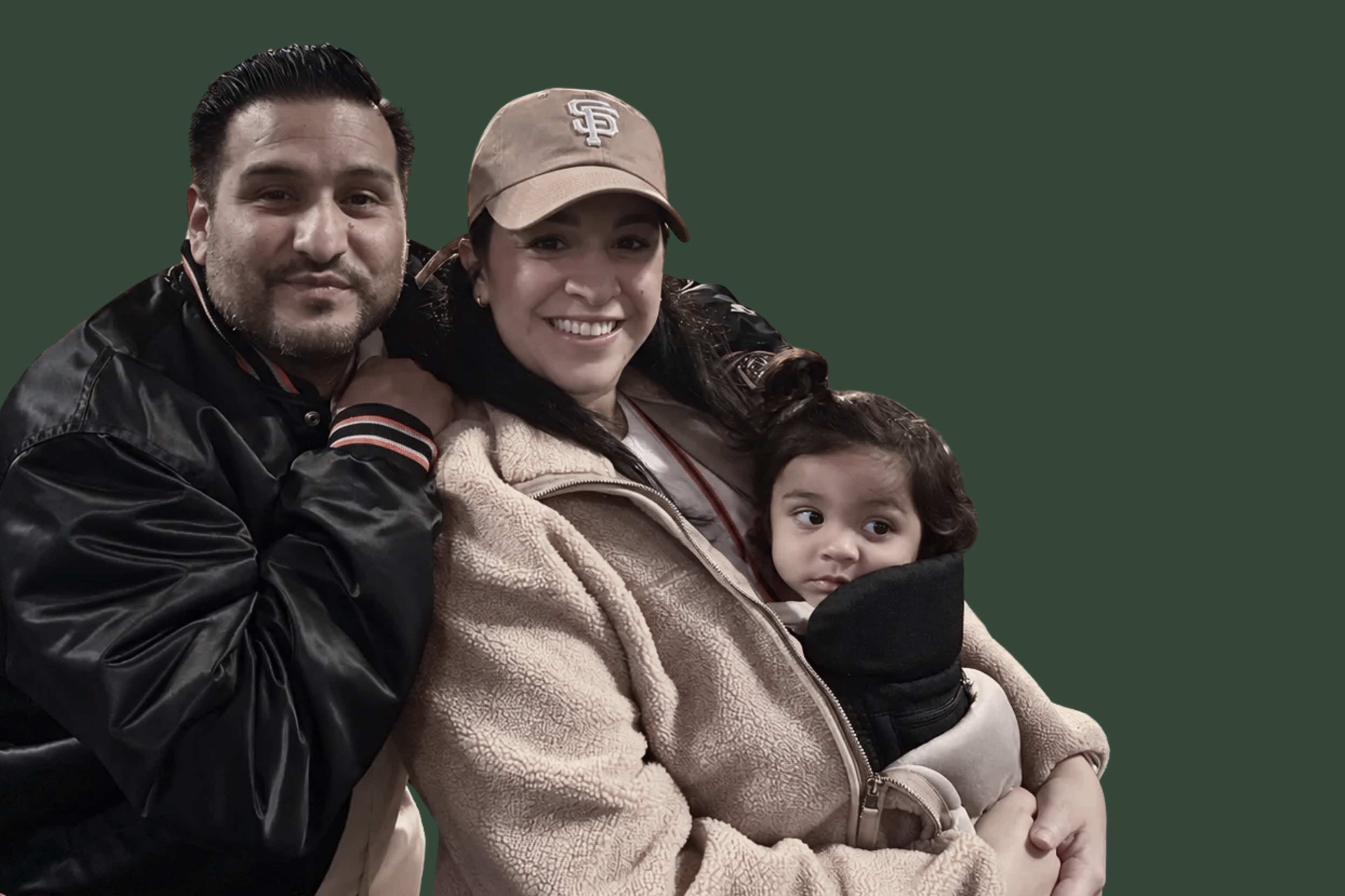 A man in a black jacket leans close to a smiling woman in a beige fleece jacket and brown cap, who is holding a young child wrapped in a black blanket.