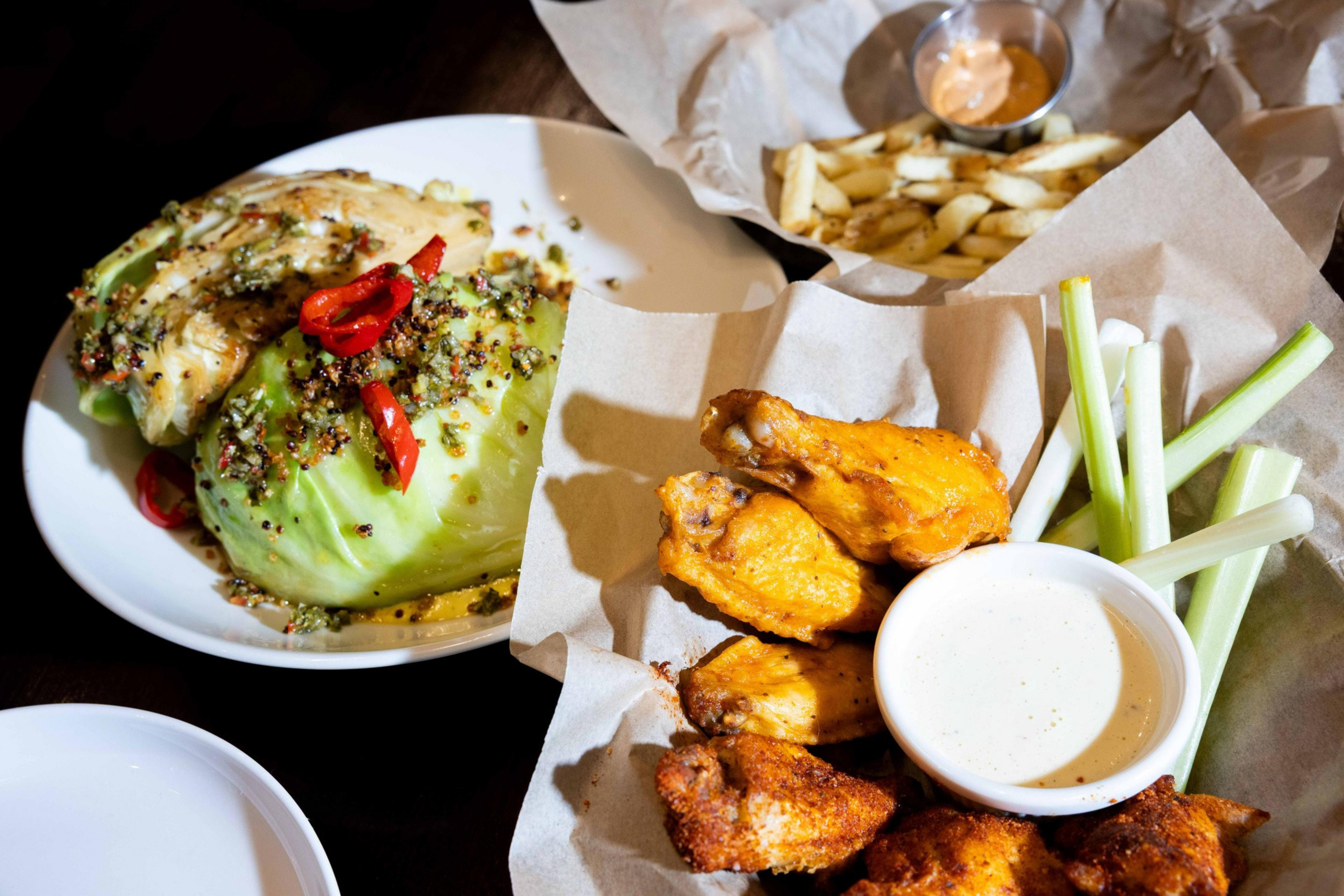 A plate of lettuce wedges with red peppers, a basket of chicken wings with a ranch dip and celery sticks, and fries with sauce in the background.