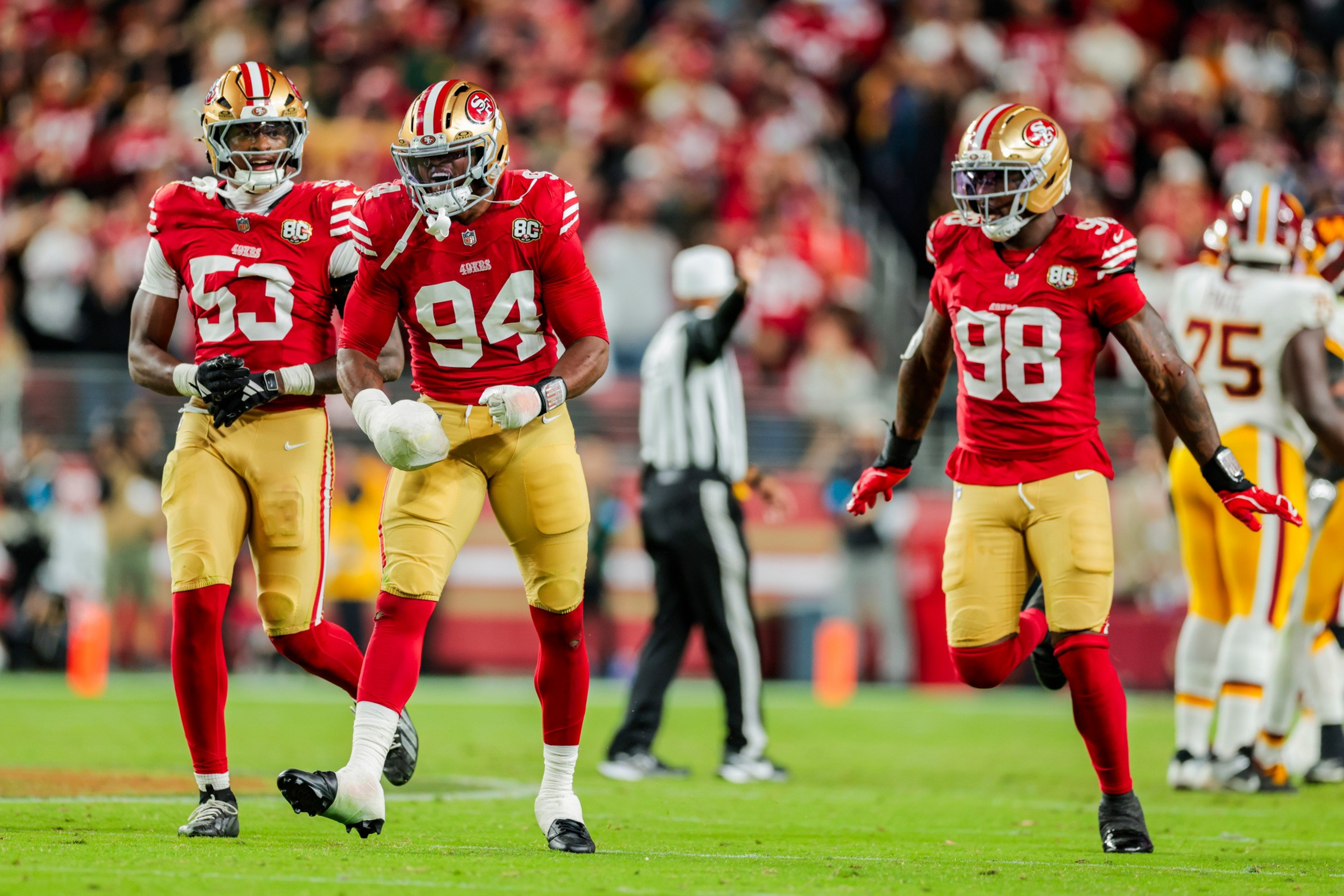 Three San Francisco 49ers players in red jerseys and gold pants celebrate on a football field with opponents and a referee in the background.