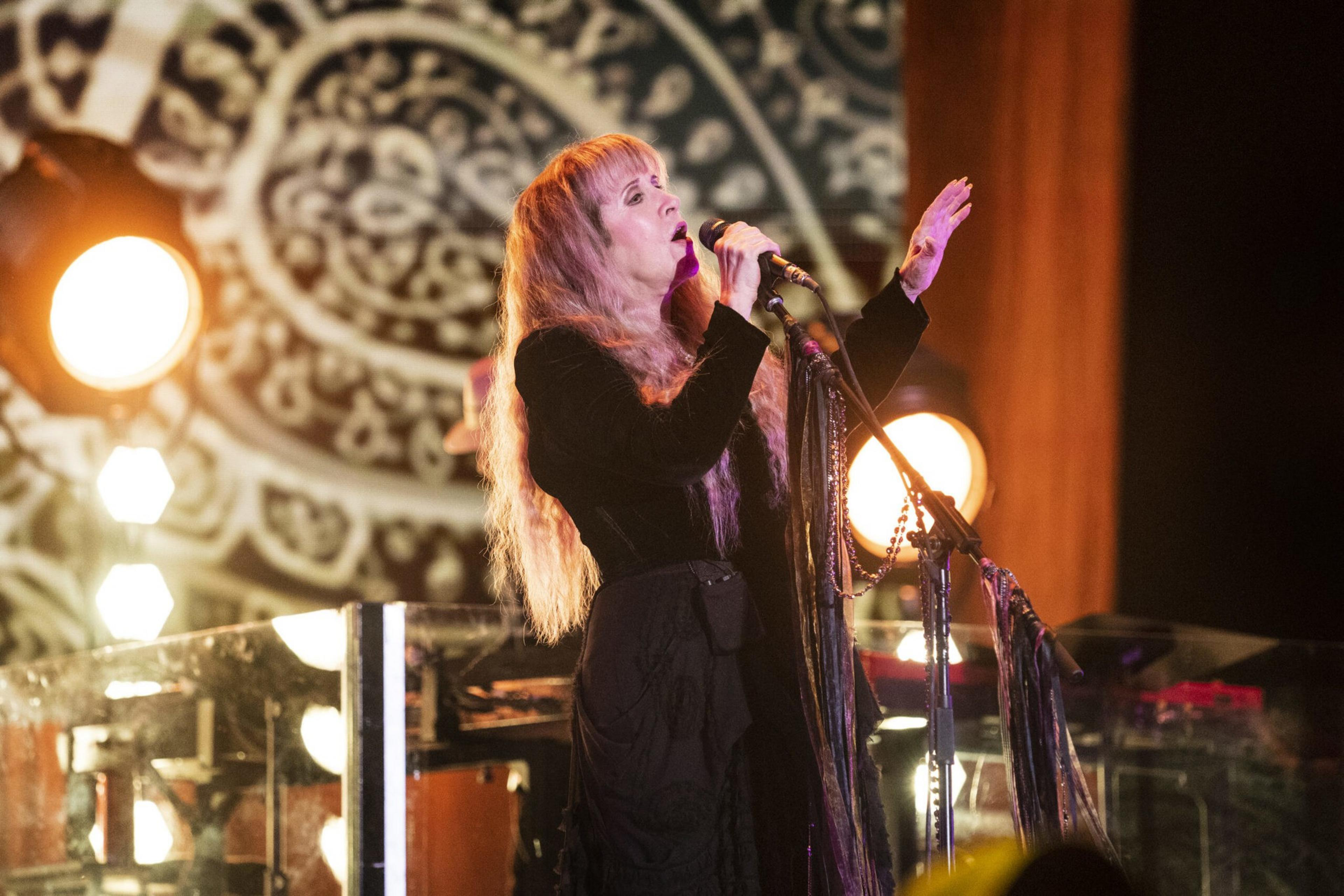 A woman with long blonde hair sings into a microphone on stage, reaching one hand upward, with bright round lights and a patterned backdrop behind her.