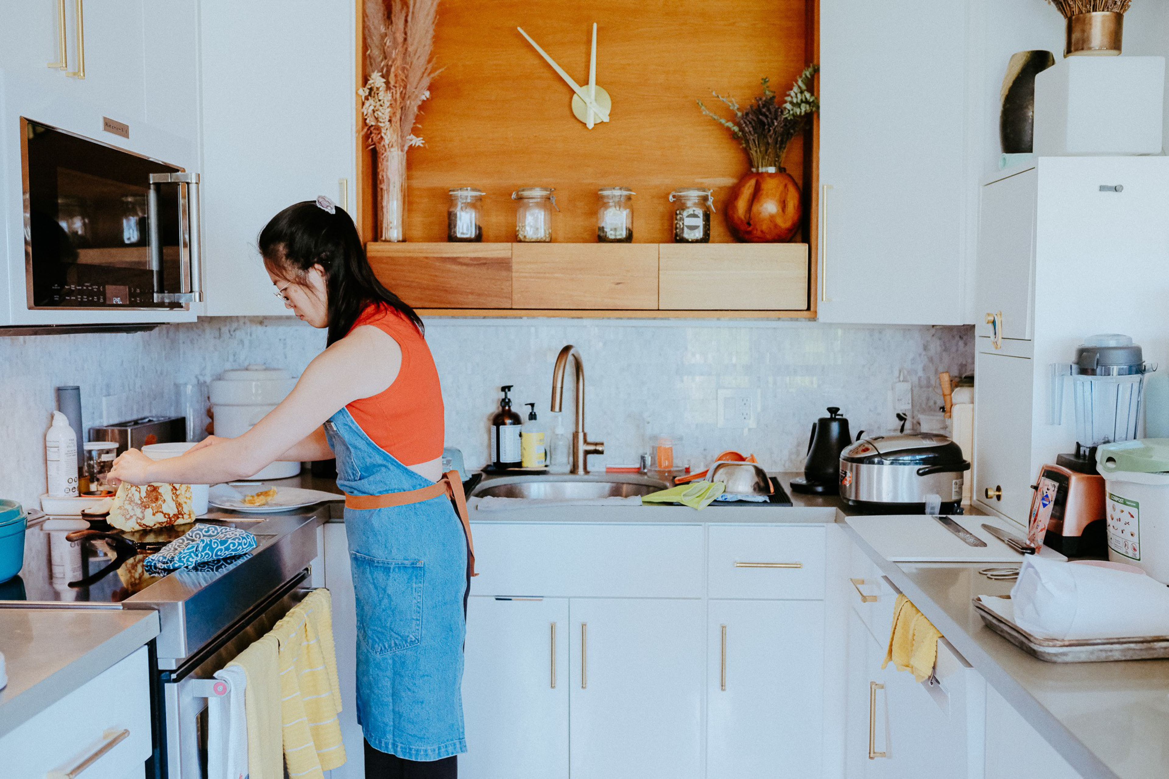 A woman in a red shirt and blue apron is preparing food on a stove in a bright kitchen with white cabinets, a wooden clock, and various kitchen appliances.