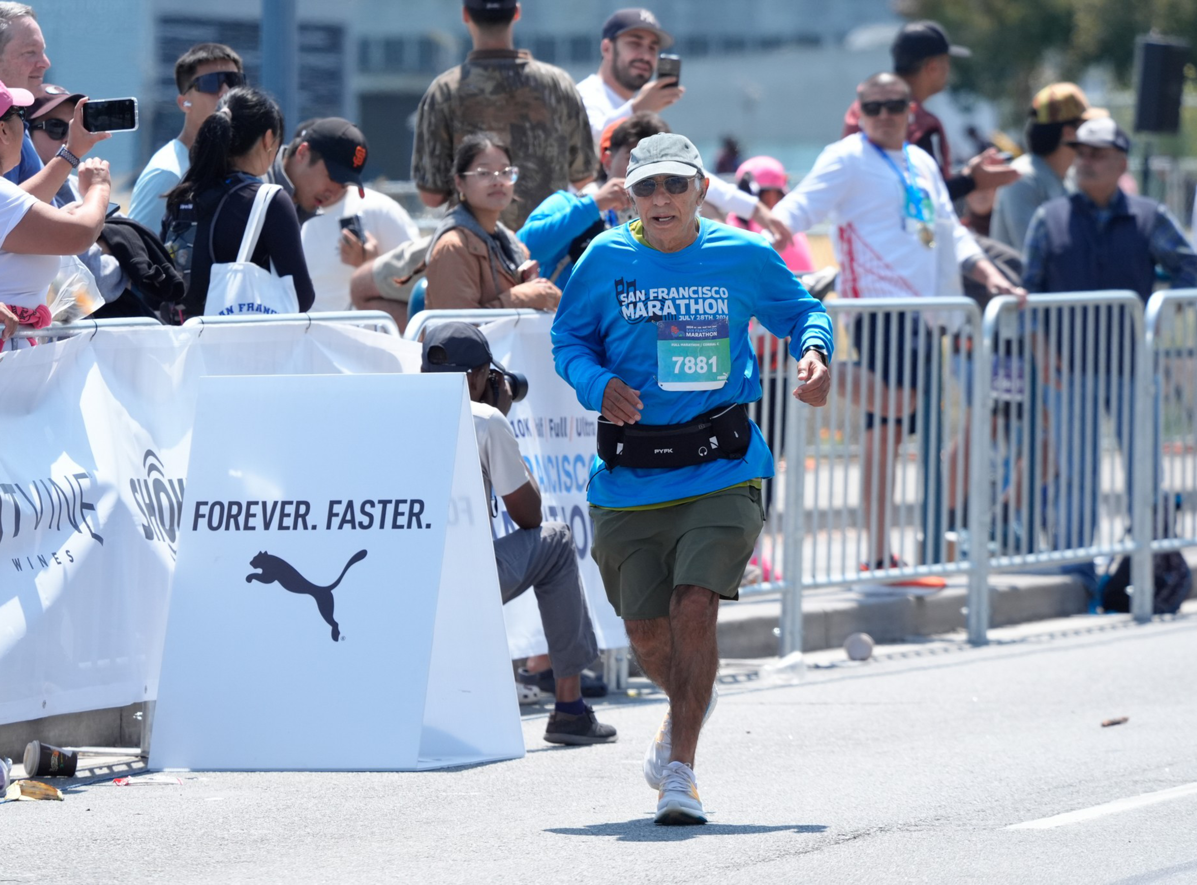A marathon runner in a blue shirt and khaki shorts nears the finish line, cheered by a crowd behind metal barriers. A sign reads &quot;FOREVER. FASTER.&quot; with the Puma logo.