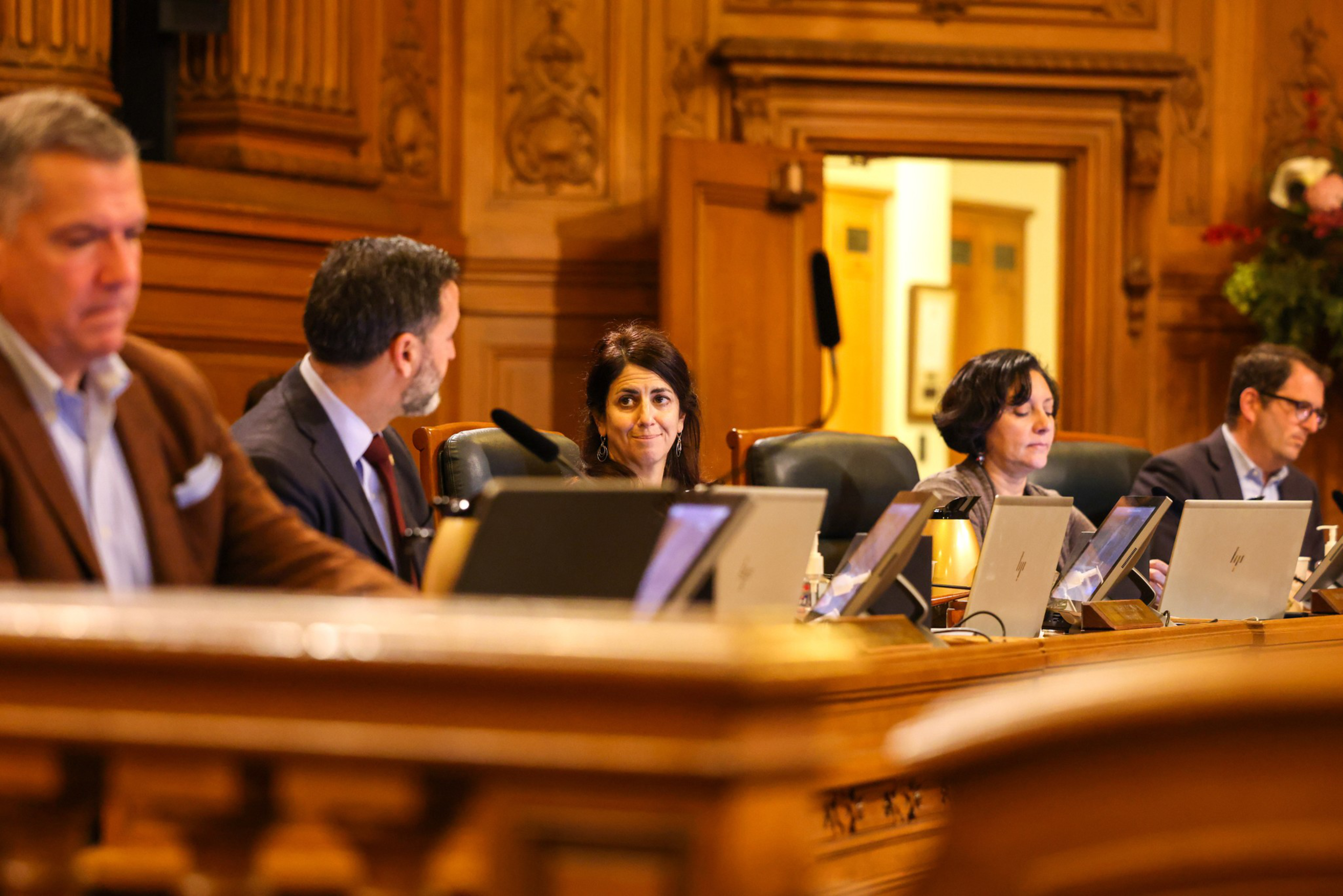 Five people, seated at a wooden panel desk with laptops, engage in discussion in a formal room with ornate wood paneling.