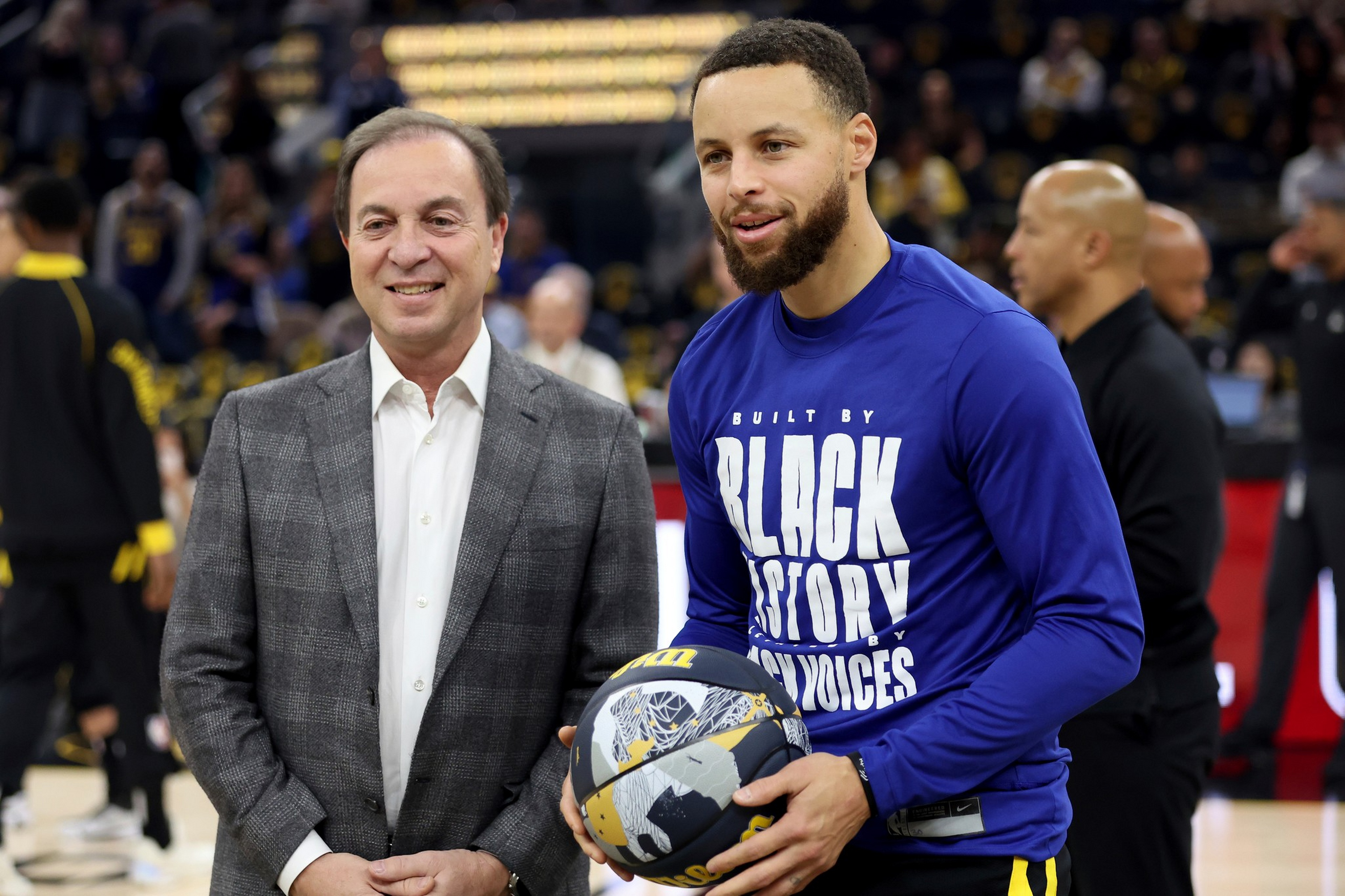 A man in a gray plaid suit stands next to a man in a blue shirt holding a basketball, both smiling on a basketball court.