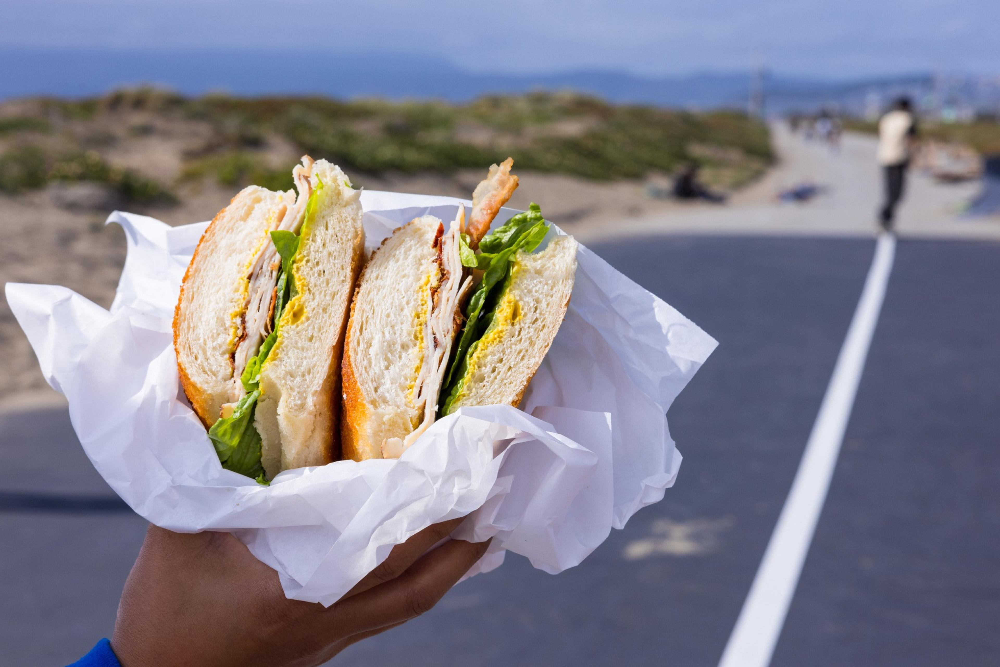 A hand holds a large sandwich with lettuce and meat, wrapped in white paper. The background shows a pathway, sand dunes, and a person in the distance.