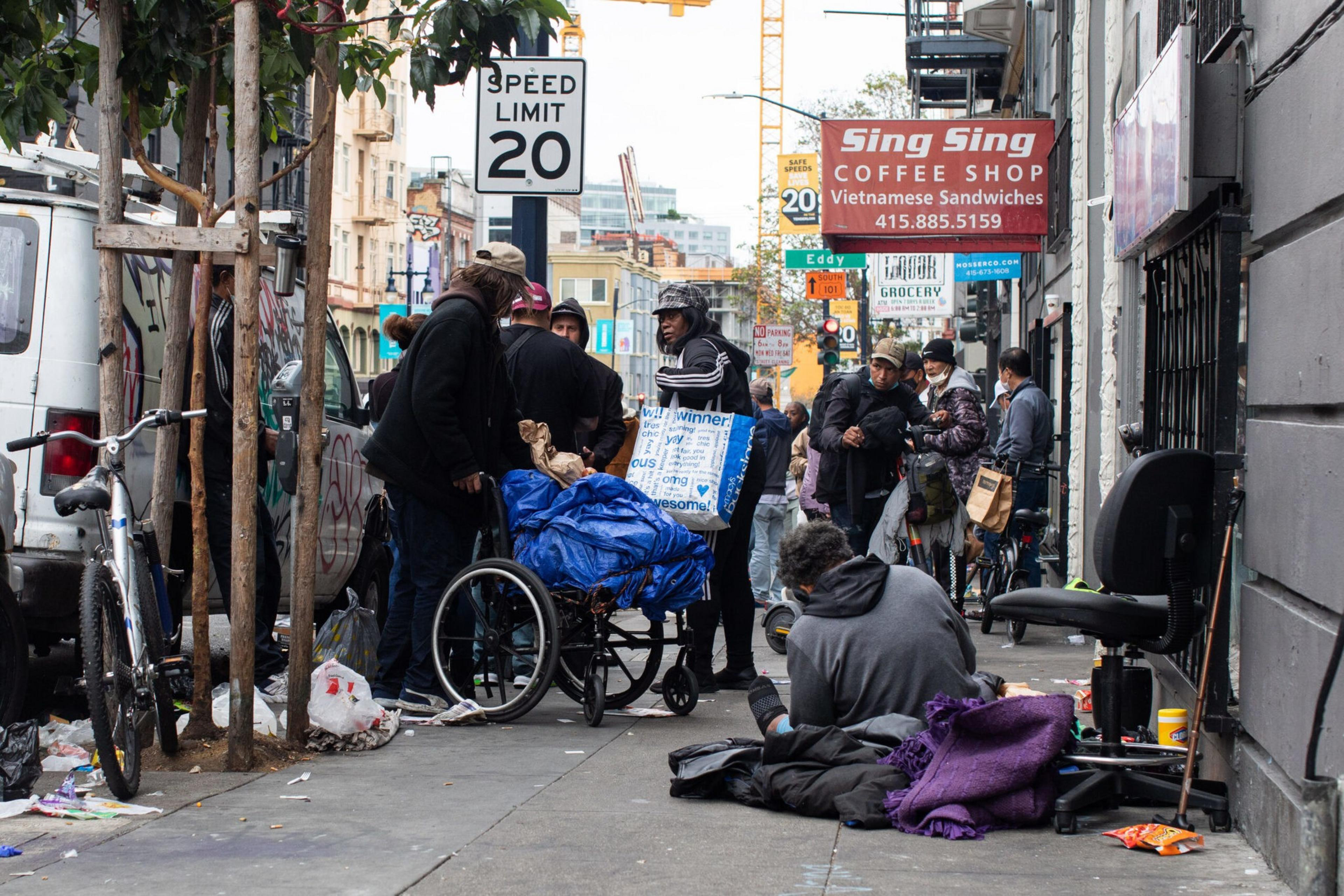 A group of people gathers on a city sidewalk near a wheelchair covered in blue fabric, with one person sitting on the ground wrapped in a purple blanket.