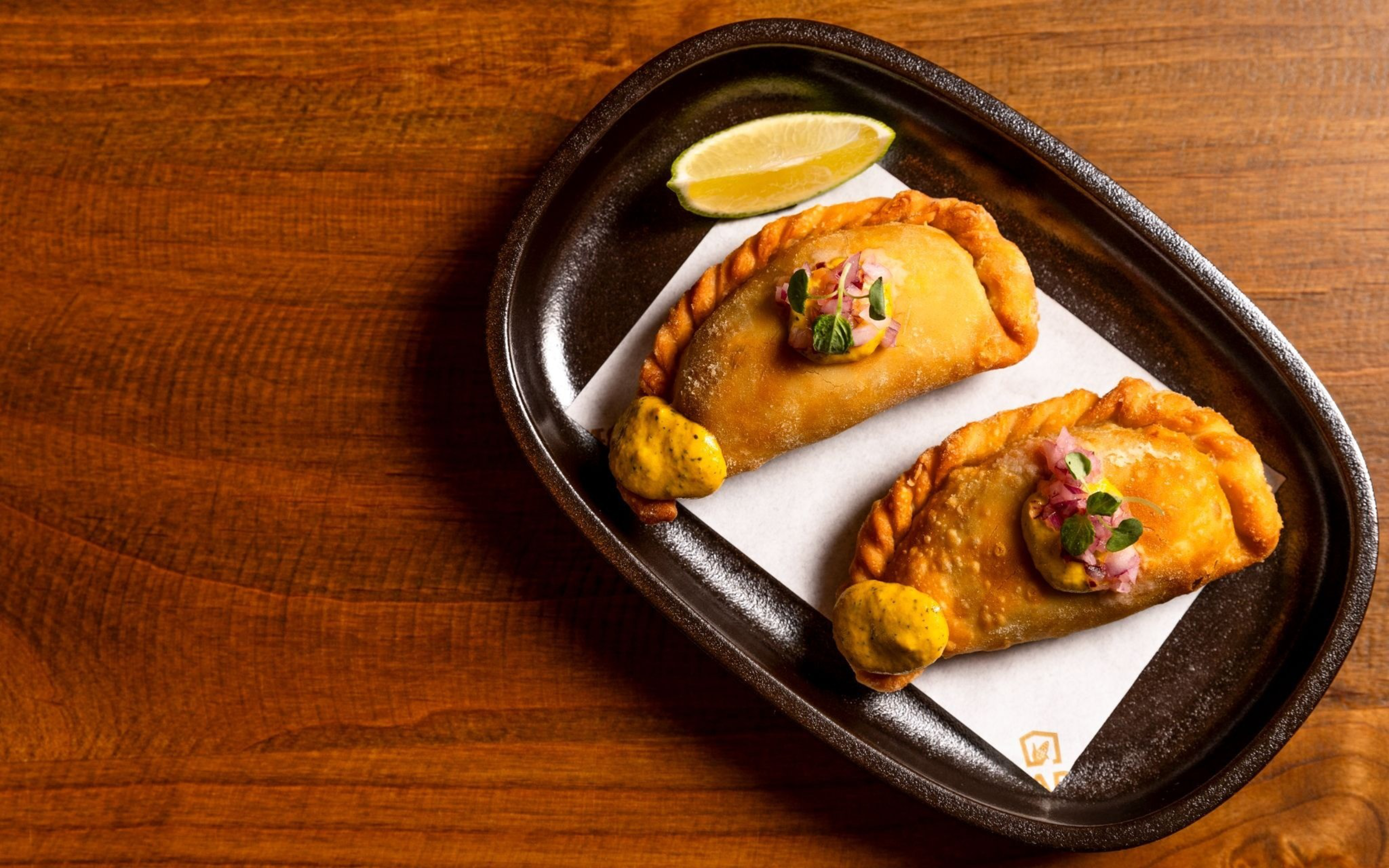 Two golden-brown empanadas topped with diced onions and herbs are served on a black plate with a lemon wedge and dollops of yellow sauce.