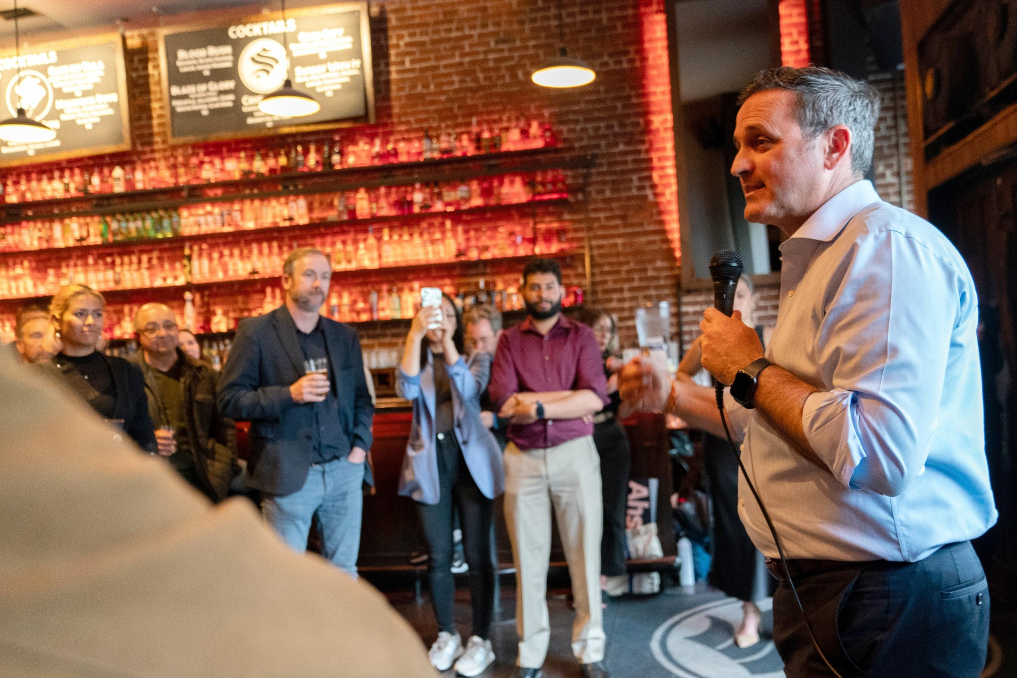 A man speaks into a microphone in a bar with attentive listeners, brick walls, a lit bar shelf, and hanging menu signs.