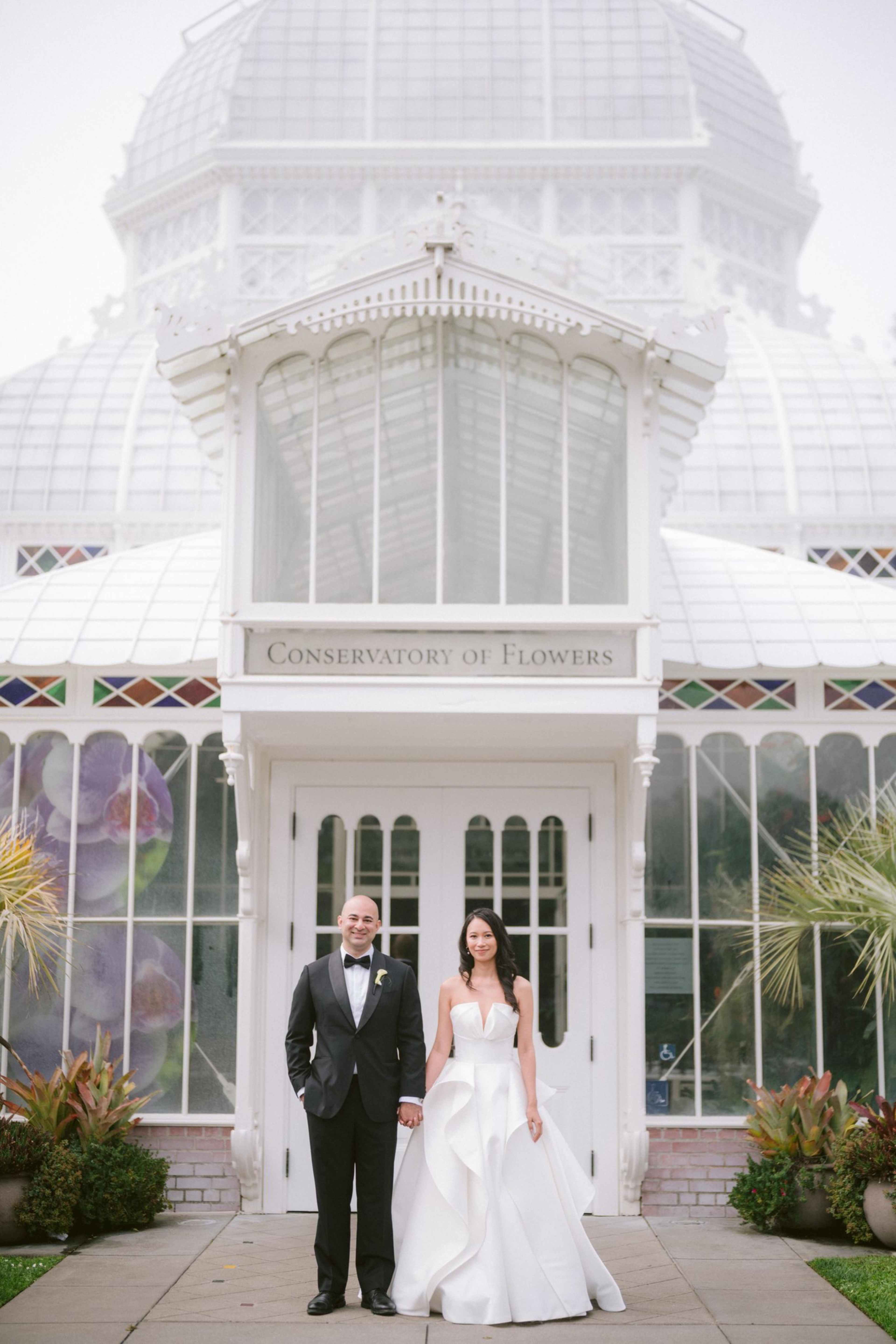 A bride in a white gown and a groom in a black tuxedo hold hands, standing in front of the white Conservatory of Flowers with glass windows and plants.