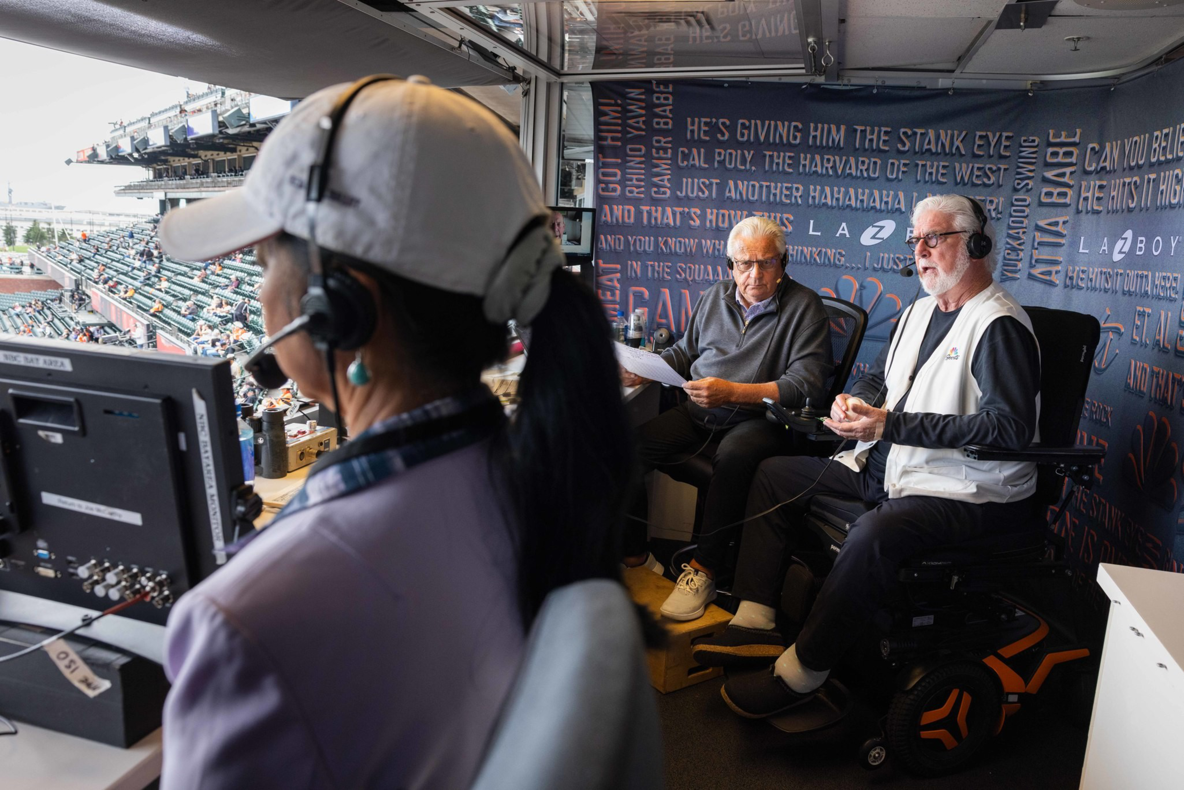 Three sports commentators wearing headsets are in a booth overlooking a stadium filled with spectators; one woman faces computer monitors while two men talk.